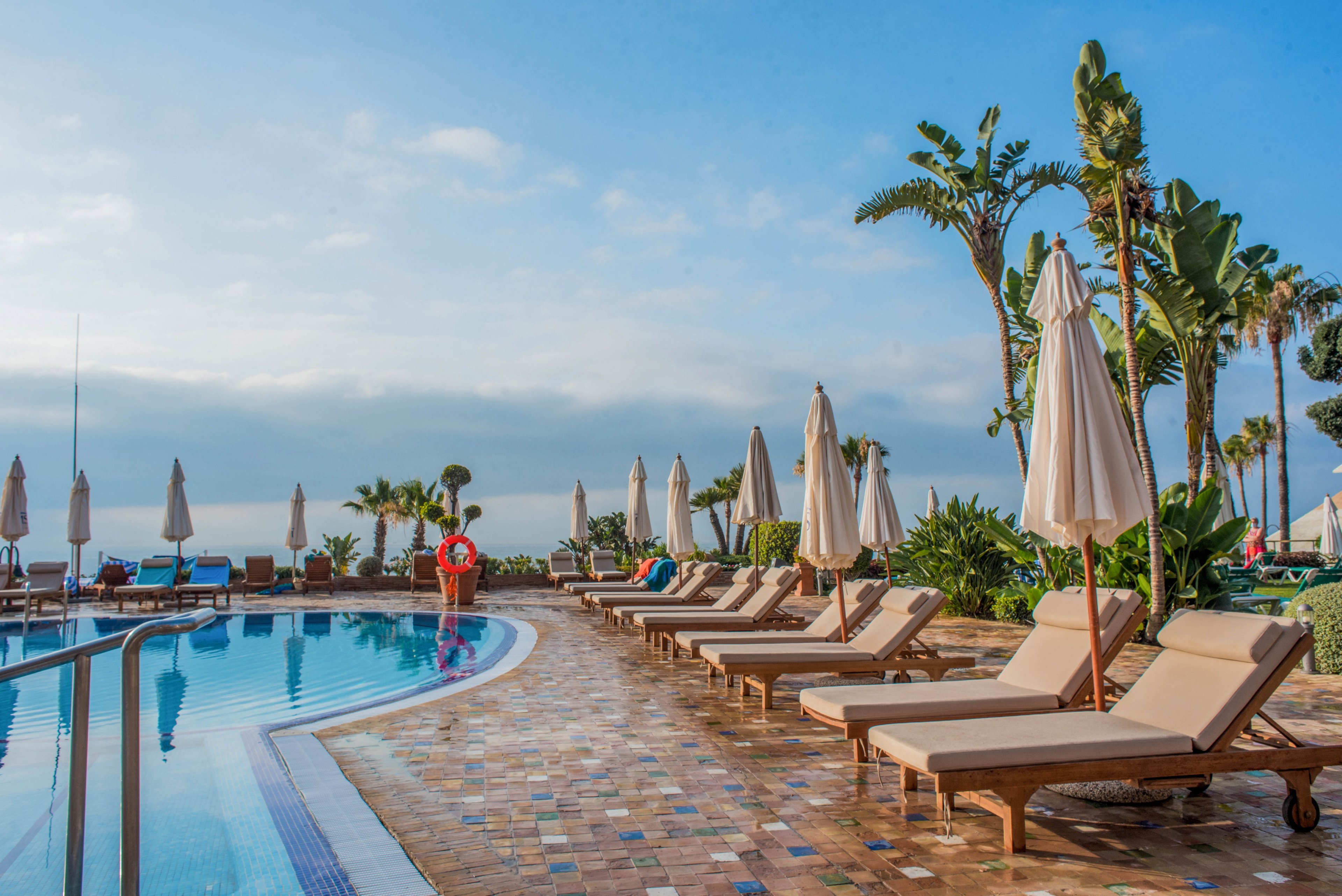 An outdoor resort swimming pool is lined with cushioned lounge chairs and closed umbrellas on a tiled deck. A single person in red stands near the pool, looking toward the water. Palm trees and lush greenery frame the scene, with the ocean and horizon visible in the background. The bright daylight and clear blue sky create a relaxed, vacation atmosphere.