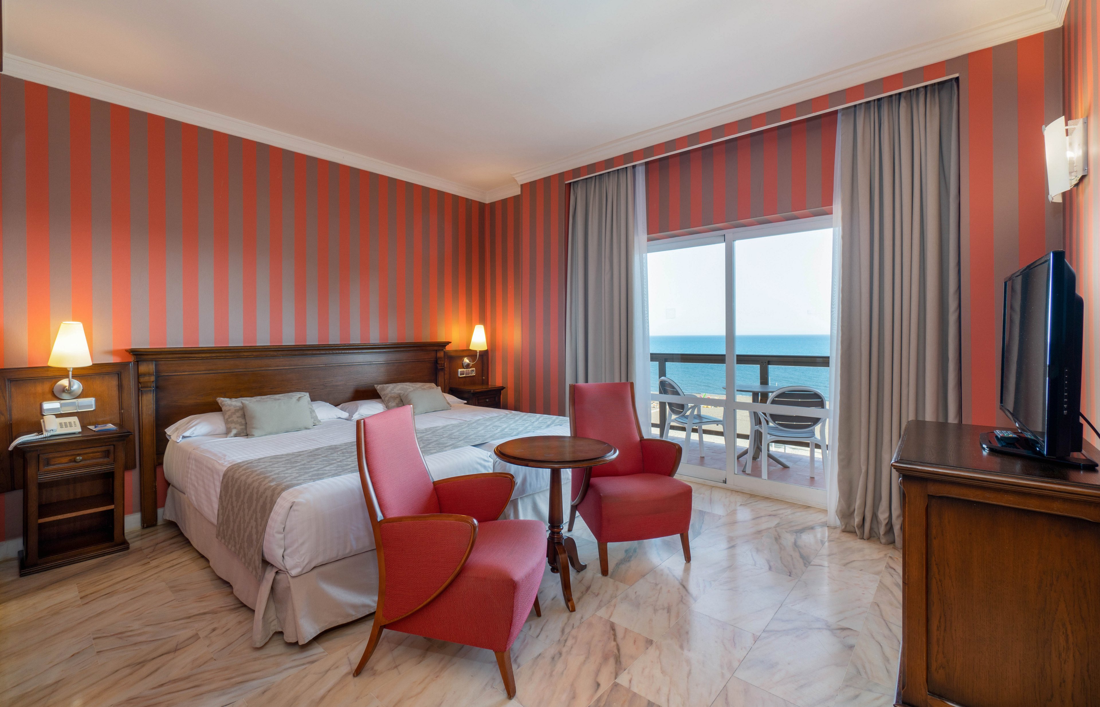 A neatly made double bed sits in a cozy hotel room with warm striped walls. Two red armchairs and a small round table create a seating area on the marble floor. Large sliding glass doors open to a balcony with chairs overlooking the ocean. Soft lighting from bedside lamps and a wall sconce adds to the relaxed, inviting atmosphere.
