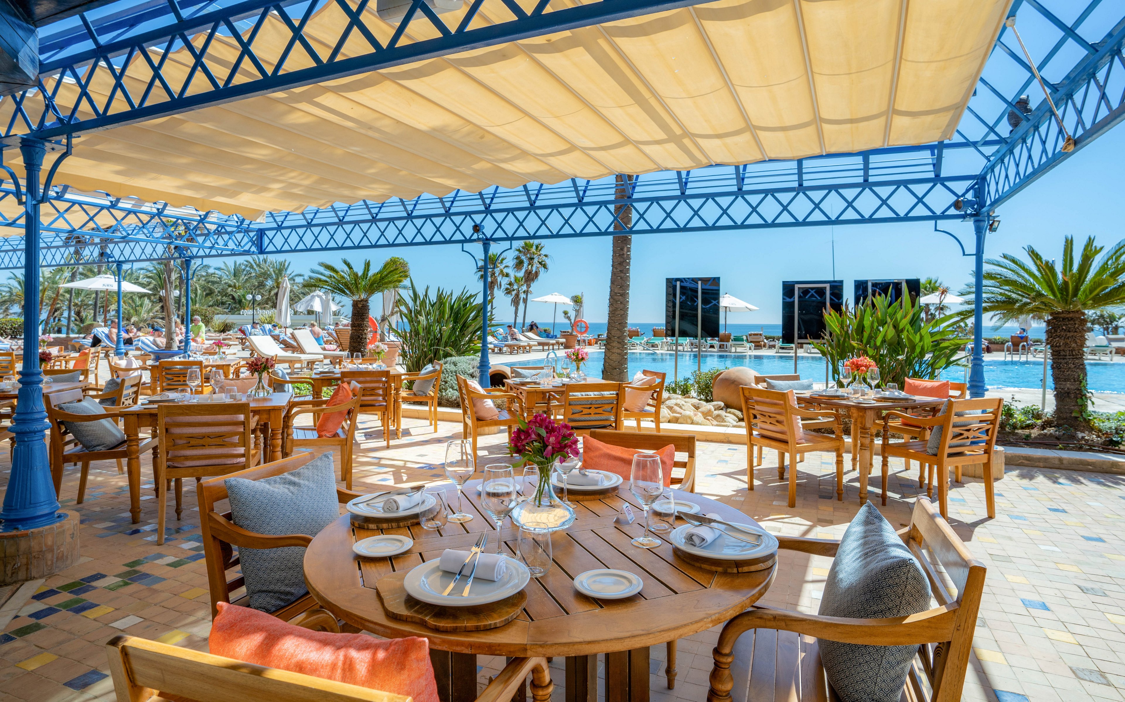 An open-air restaurant terrace features wooden tables set with plates, glasses, and cutlery. Cushioned chairs and colorful pillows create a relaxed dining atmosphere under a large canopy. In the background, guests lounge by a bright blue swimming pool surrounded by palm trees. The sunny weather and clear sky emphasize a tropical, vacation resort setting.