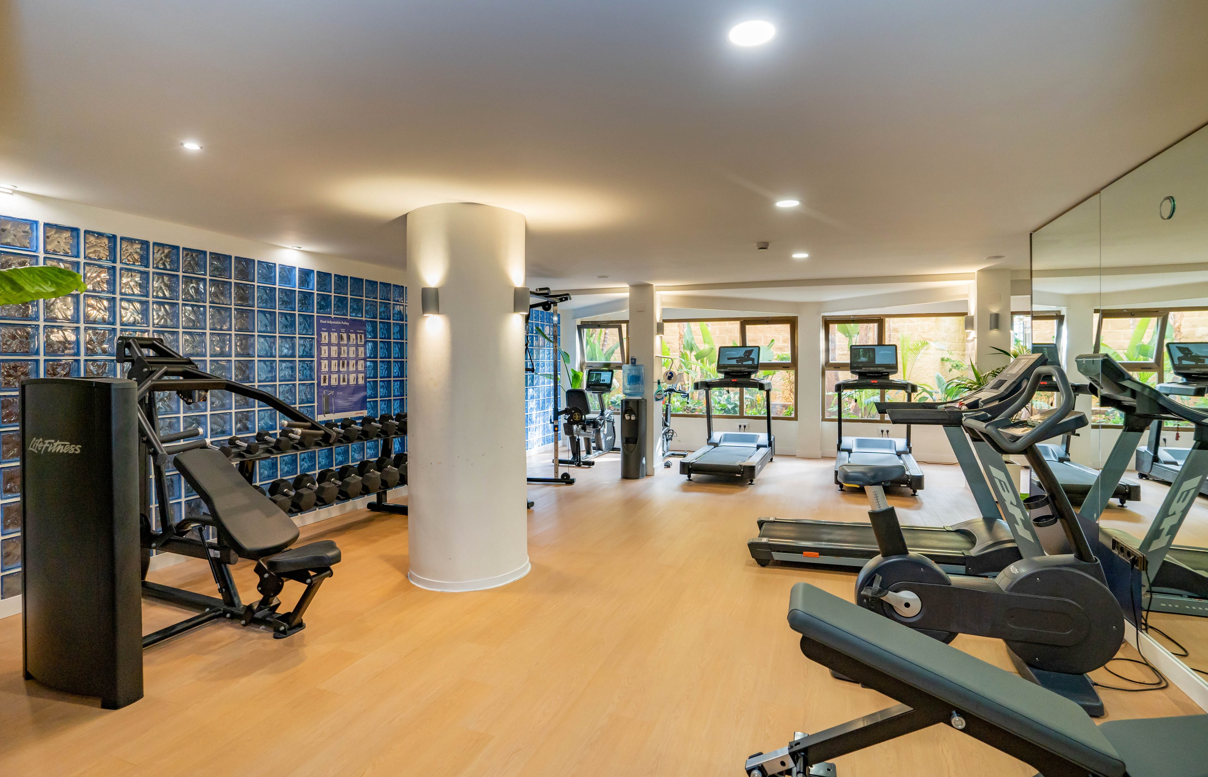 A bright, modern gym features various exercise machines neatly arranged on a light wood floor. Treadmills, stationary bikes, and strength-training equipment face large windows that overlook greenery outside. Recessed ceiling lights and mirrored walls enhance the spacious, clean atmosphere. The setting suggests a hotel or residential fitness center designed for everyday workouts and wellness.