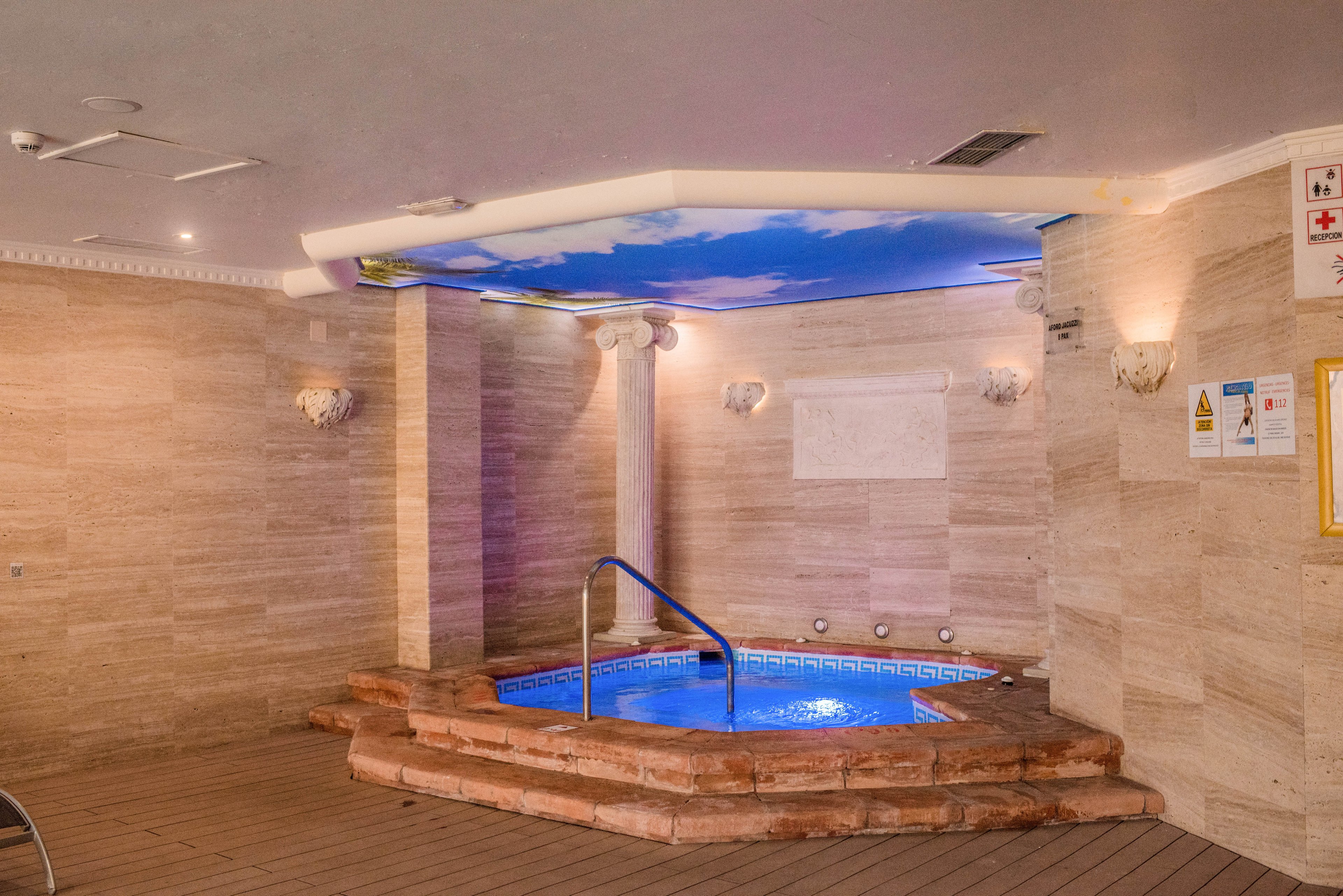 An indoor spa area features a small circular jacuzzi pool surrounded by stone tiling. The hot tub is set into a raised brick platform with a metal handrail for safe entry. Soft wall sconces and recessed lighting create a warm, relaxing atmosphere, while the ceiling displays a sky mural. Safety and instruction signs are mounted on the wall near the pool.