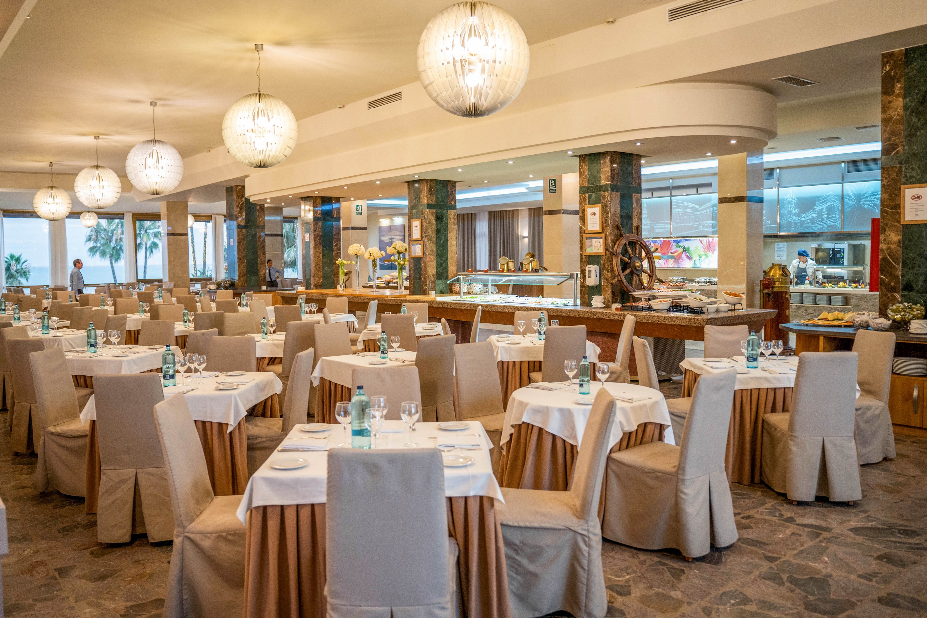Spacious restaurant dining room with neatly arranged round tables covered in white tablecloths and beige chair covers. The setting appears ready for guests, with glassware, plates, and bottled water placed on each table. Large spherical pendant lights hang from the ceiling, illuminating the stone floor and buffet counters in the background. The overall atmosphere is formal yet inviting, suitable for events or hotel dining.