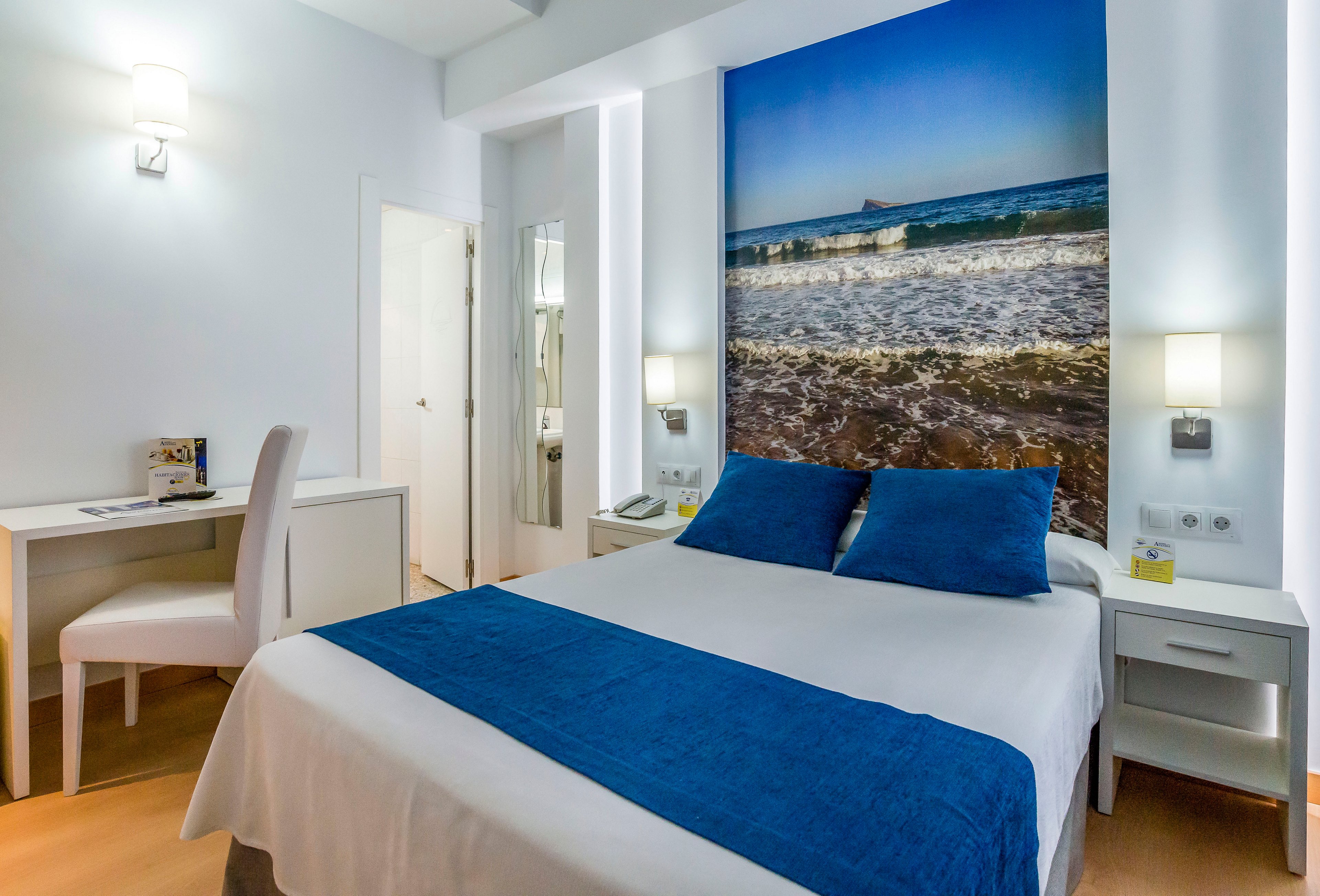 A contemporary hotel bedroom features a neatly made double bed with white linens and blue accents. Large wall art above the headboard shows a vivid ocean scene with waves and a distant boat. The room includes bedside tables with lamps, a telephone, and a small desk with a chair near the doorway. Clean white walls and light wood flooring create a fresh, minimalist atmosphere.