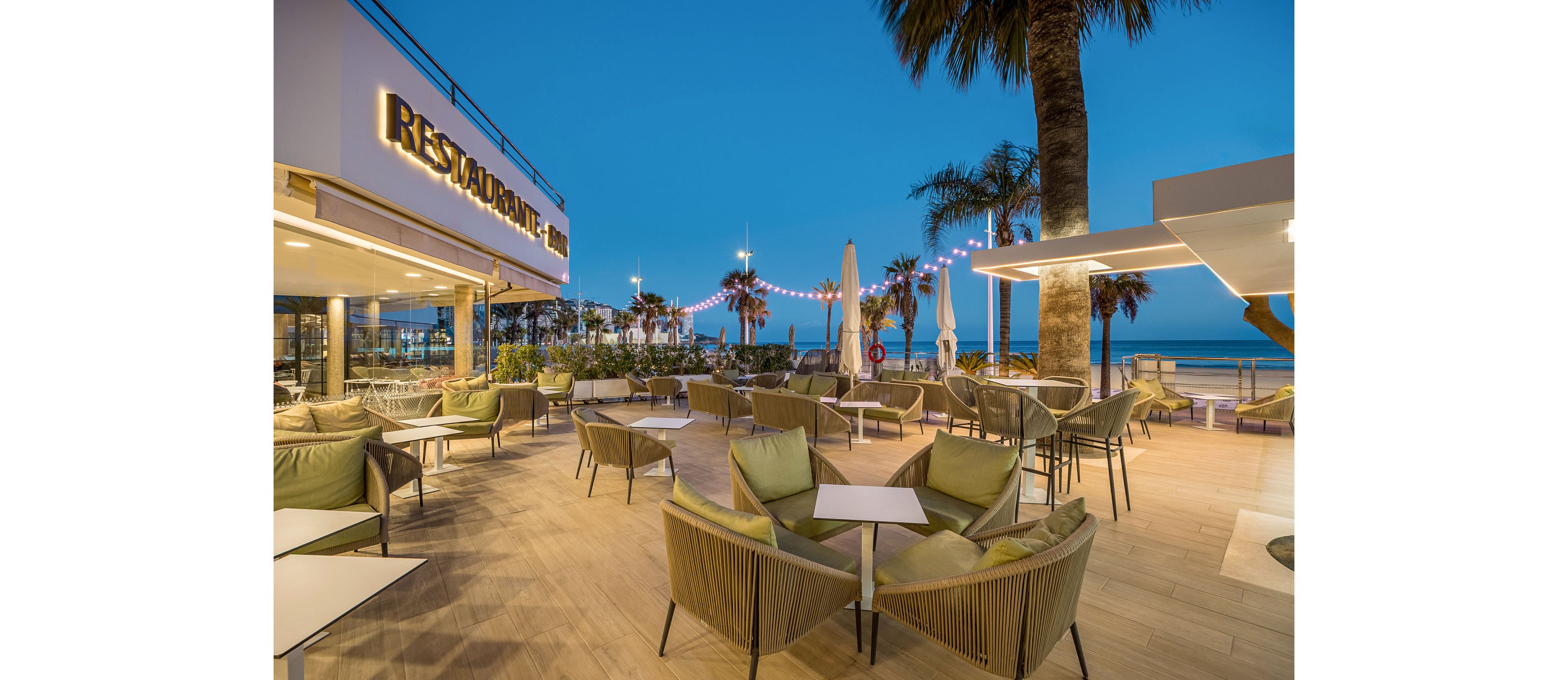 An elegant outdoor restaurant terrace overlooks a beach at dusk, with contemporary lounge chairs and tables neatly arranged. String lights and warm architectural lighting create a cozy atmosphere against the deep blue evening sky. Palm trees and large umbrellas frame the view toward the sea, suggesting a relaxed coastal setting. The scene is calm and inviting, with no people present and a focus on the stylish design of the space.