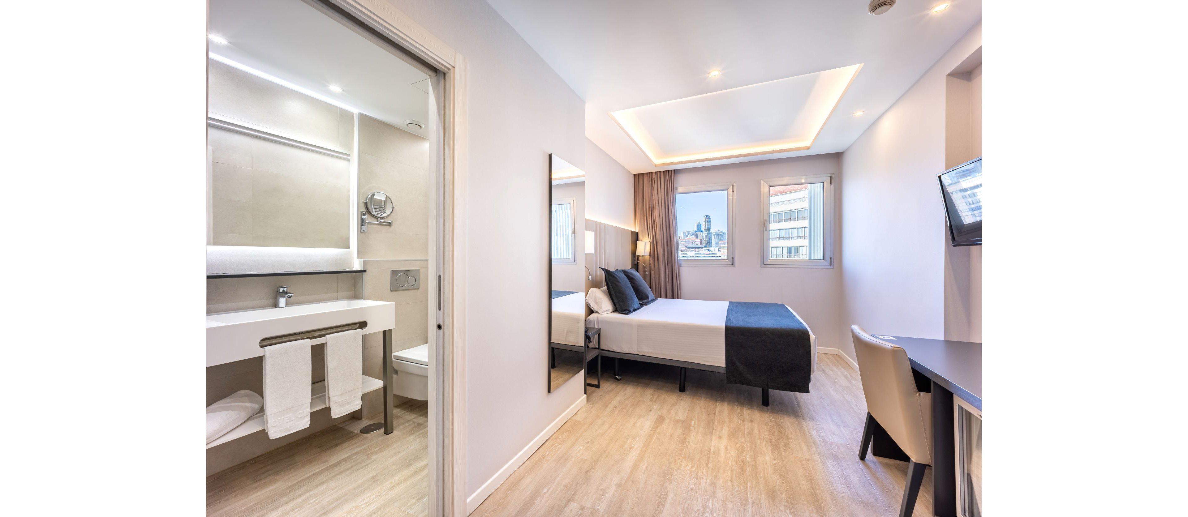 Bright contemporary hotel room featuring a neatly made double bed with black and white linens. A sleek desk and chair face a wall-mounted television, while large windows reveal a cityscape outside. The adjoining bathroom includes a vanity with towels, mirror, and shower area, all in a clean minimalist style. Light wood flooring and soft recessed ceiling lighting create an inviting, comfortable atmosphere.