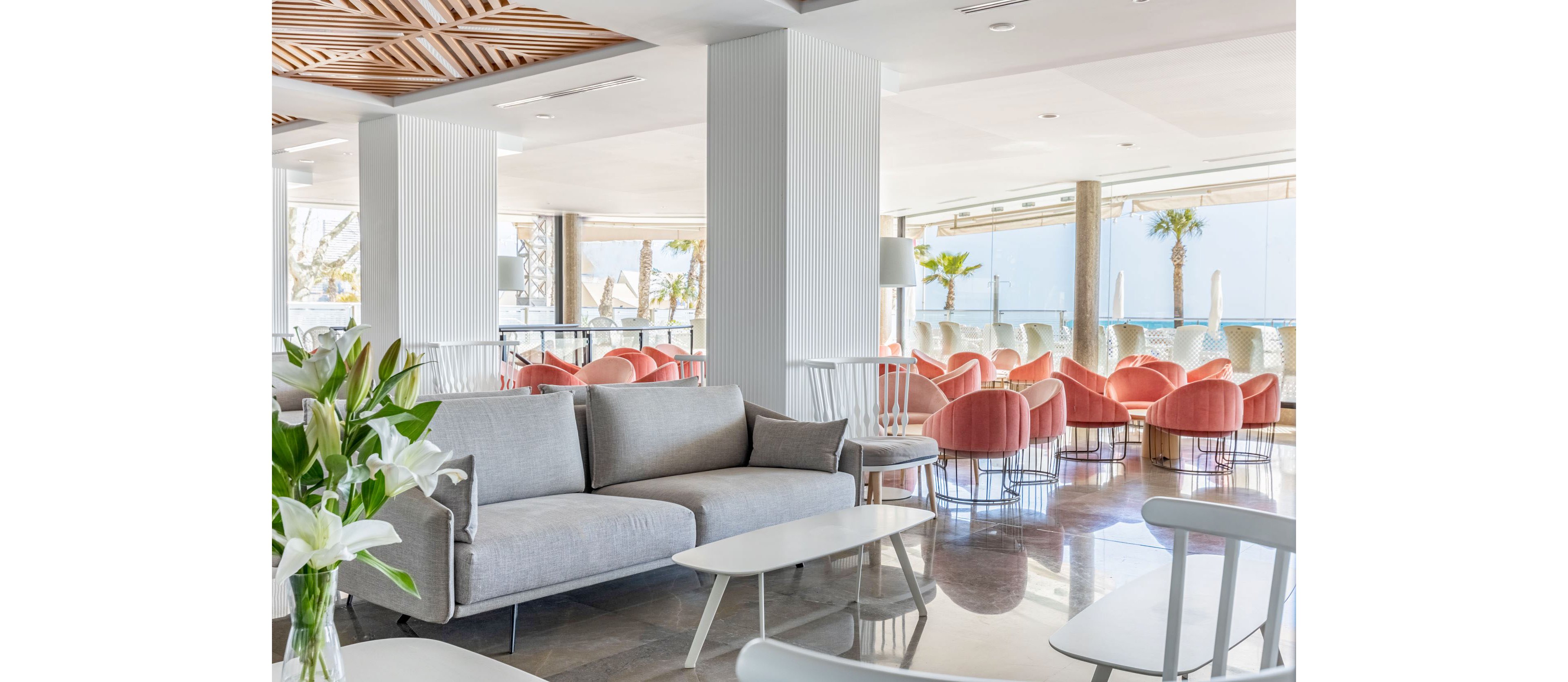 Contemporary lounge area features light gray sofas and white tables in the foreground. Soft pink upholstered chairs are arranged in groups near large floor-to-ceiling windows. Natural light floods the space, highlighting the polished floor and wooden ceiling details. The setting overlooks palm trees and a distant ocean, creating a relaxed, airy atmosphere.
