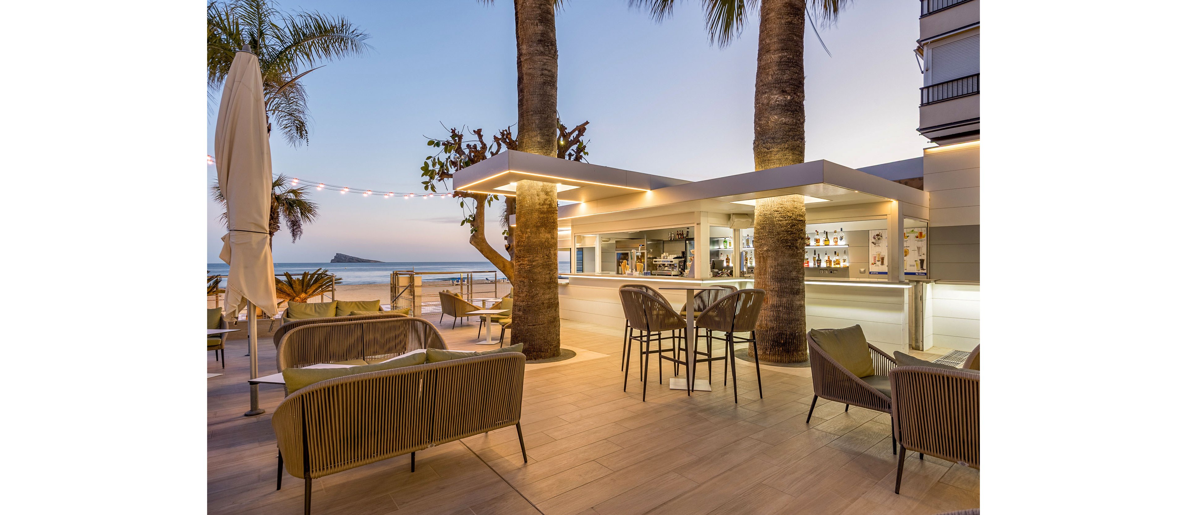 An open-air bar terrace is set between tall palm trees overlooking a calm beachfront at dusk. Contemporary chairs and high stools surround the sleek white bar counter, creating a relaxed social space. Soft string lights and warm interior lighting illuminate the scene as the sky transitions from day to evening. The ocean and a distant island are visible in the background, enhancing the vacation atmosphere.