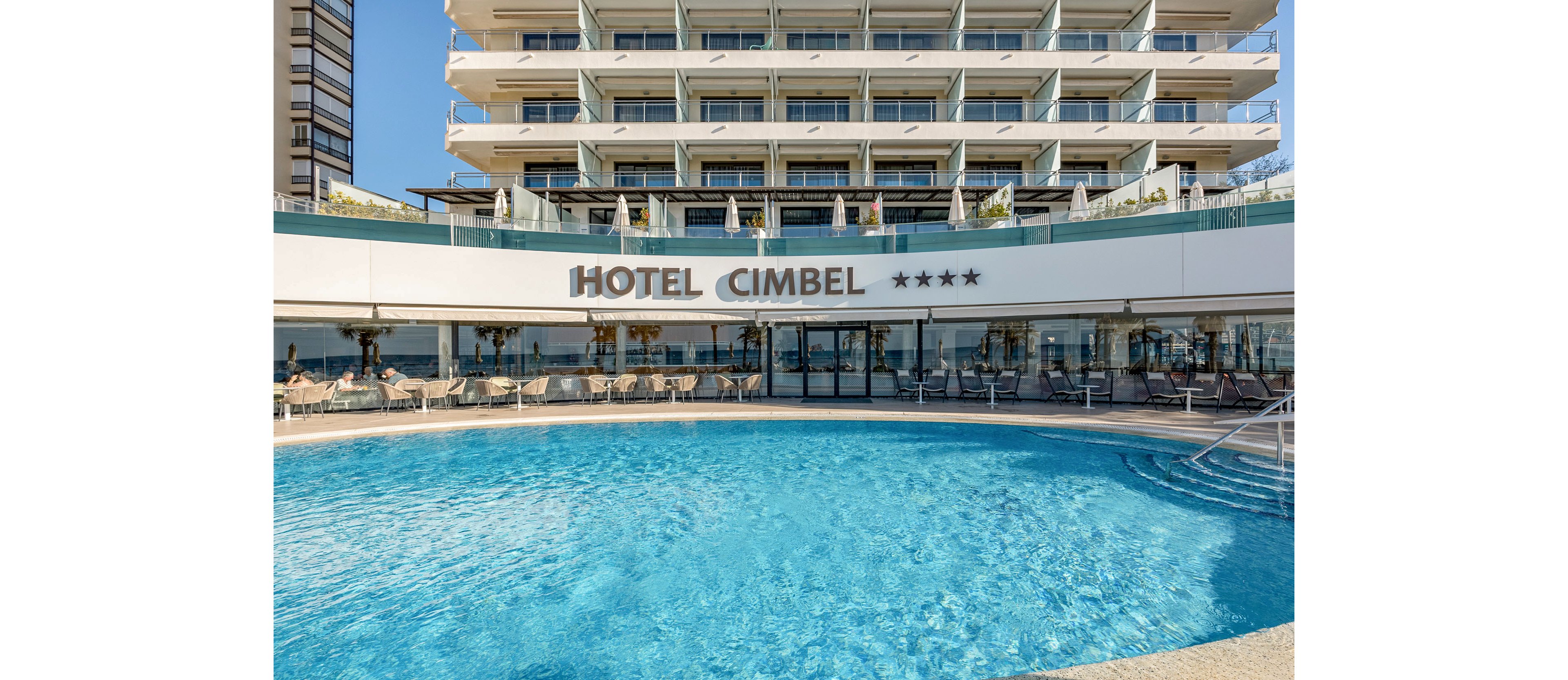 A large outdoor swimming pool sits in front of the Hotel Cimbel building. The multi-story hotel features balconies facing the pool and the sea beyond. Sun loungers and umbrellas line the poolside terrace, suggesting a relaxing vacation setting. The clear blue water and bright daylight create a sunny, coastal atmosphere, and four stars are displayed under the hotel name.