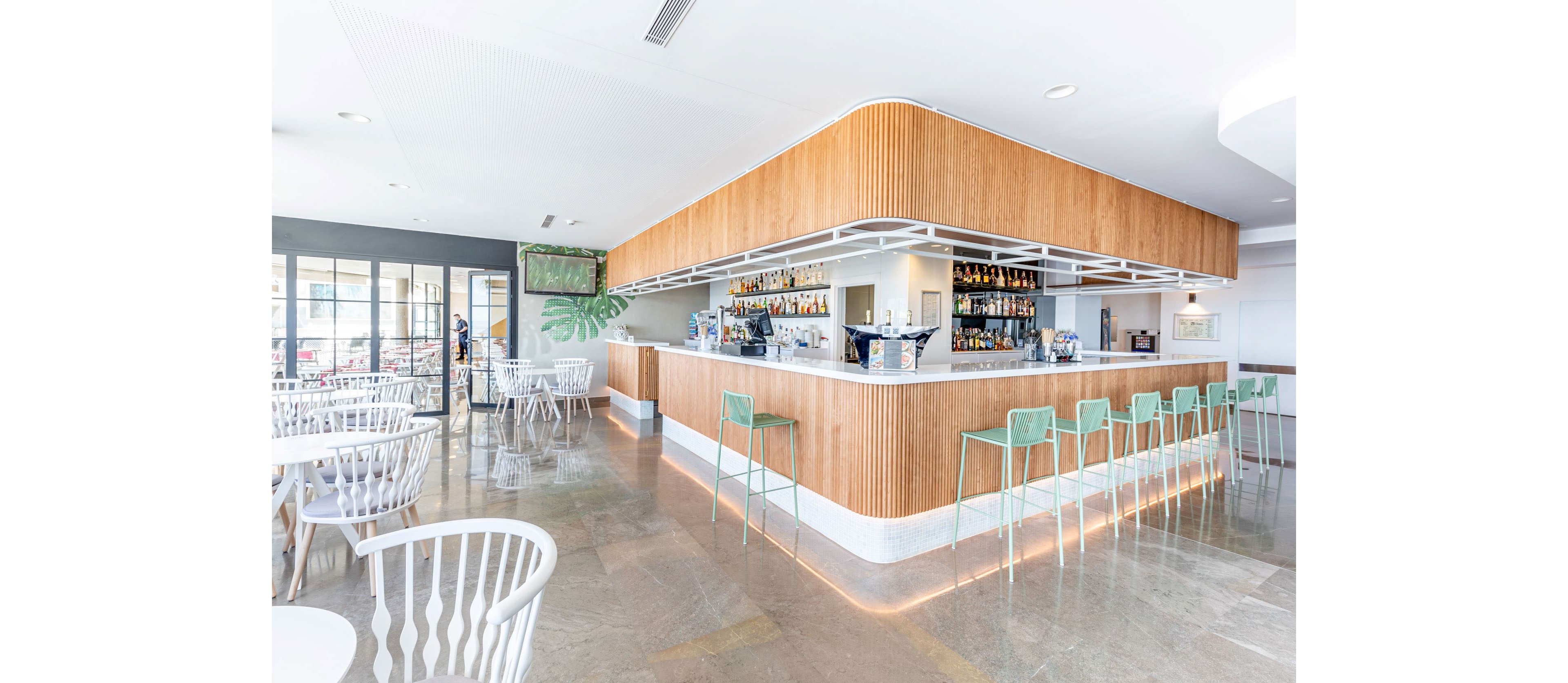 Contemporary bar interior with a long counter and backlit shelves of bottles is shown in a bright, open space. Mint-green bar stools line the counter, while white chairs and round tables fill the surrounding seating area. Large windows bring in natural light, reflecting off the polished floor and highlighting the clean, minimalist design. The overall atmosphere feels airy, relaxed, and inviting for guests.