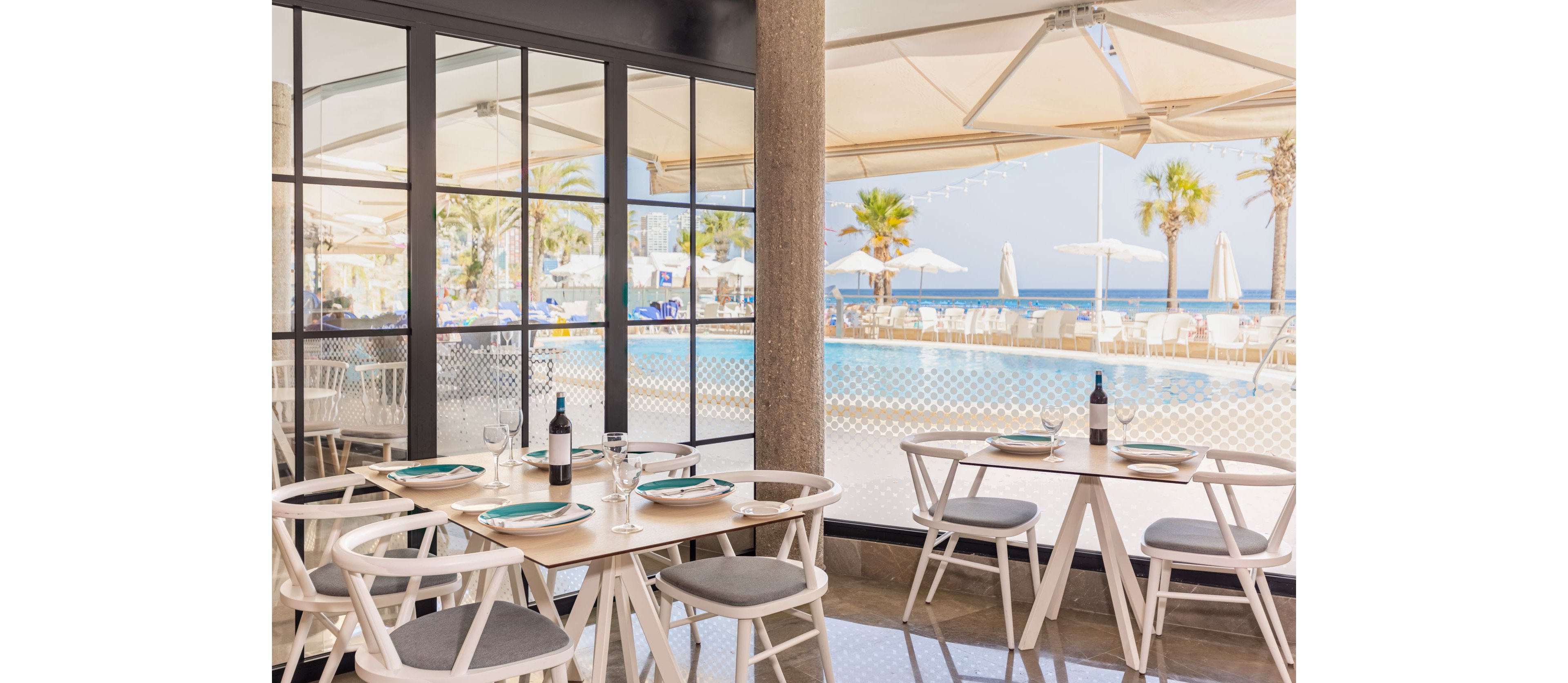 A modern seaside restaurant terrace features light wooden tables and white chairs arranged neatly near large glass windows. Wine bottles and glasses are set on the tables, suggesting a relaxed dining experience. Outside, a swimming pool, palm trees, and sun umbrellas frame the view toward the ocean under a clear blue sky. The overall scene feels airy, inviting, and ideal for vacation leisure.