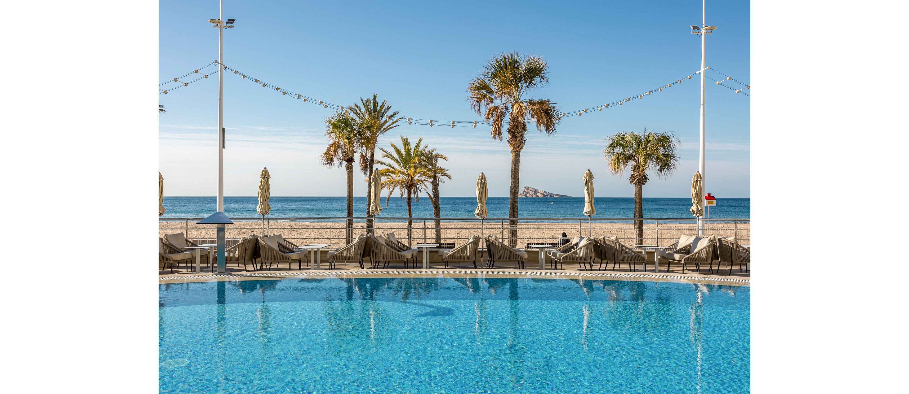 A clear blue swimming pool sits in the foreground of a coastal resort scene. Sun loungers and closed umbrellas line the poolside, facing a sandy beach and calm ocean waters. Tall palm trees and decorative string lights add a relaxed, vacation atmosphere. In the distance, a small rocky island rises from the sea under a bright, clear sky.