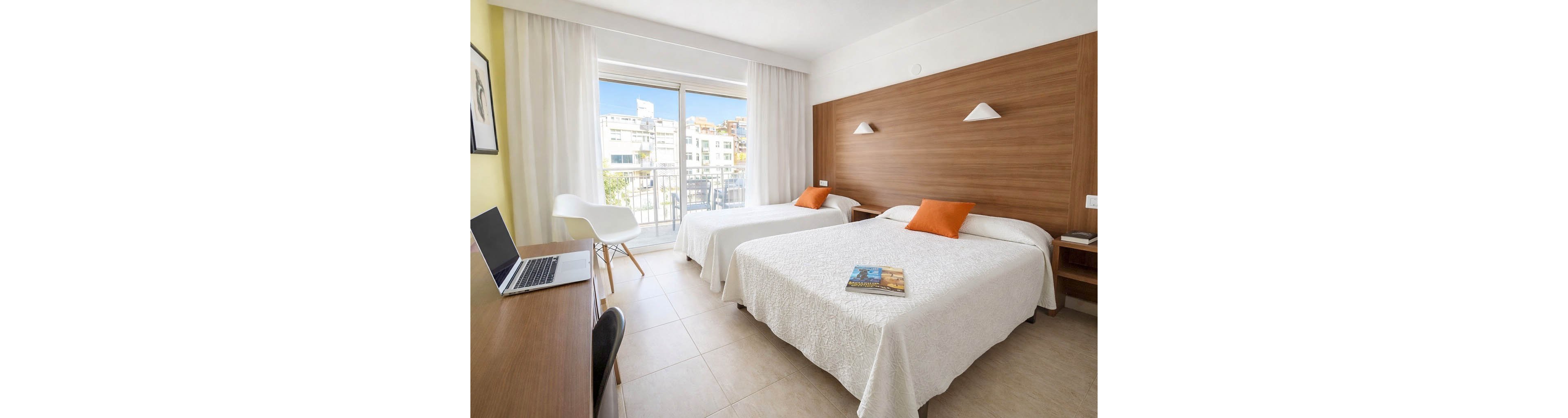 A bright hotel room features two neatly made twin beds with white bedspreads and orange accent pillows. A sliding glass door opens to a balcony with a city view, allowing natural light to fill the space. A wooden desk with an open laptop and a modern white chair sits to the left, while a wooden headboard wall and bedside table complete the minimalist decor. A book or magazine lies on one of the beds, adding a casual, welcoming touch.