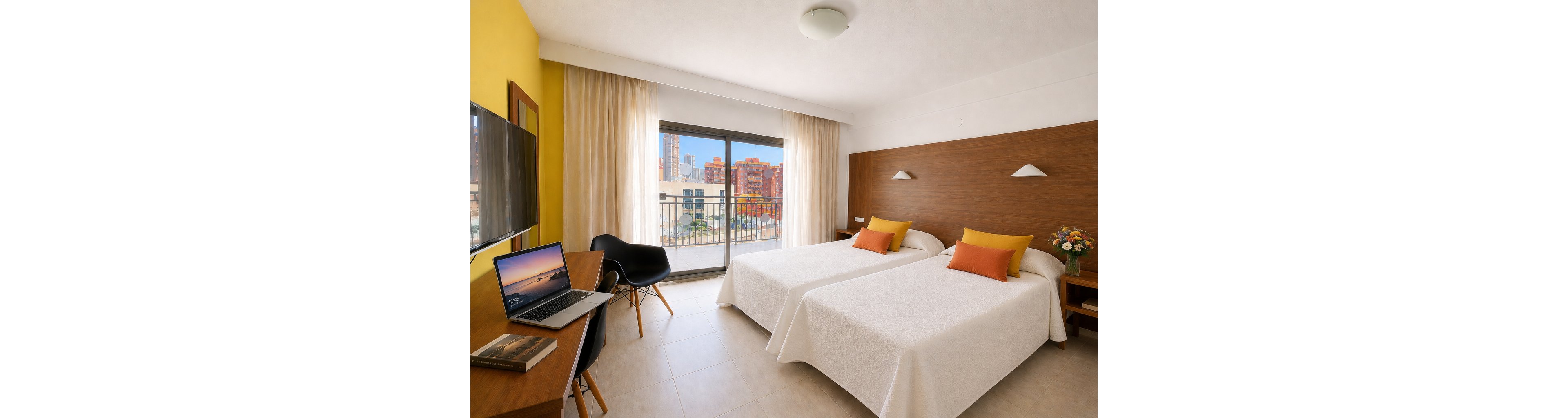 A modern hotel room features two neatly made twin beds with white linens and orange accent pillows. The space includes a wooden desk with a laptop, books, and a flat-screen TV mounted on the wall. Large sliding glass doors open to a balcony overlooking a cityscape of colorful buildings. Neutral tones, natural light, and simple decor create a clean and inviting atmosphere.
