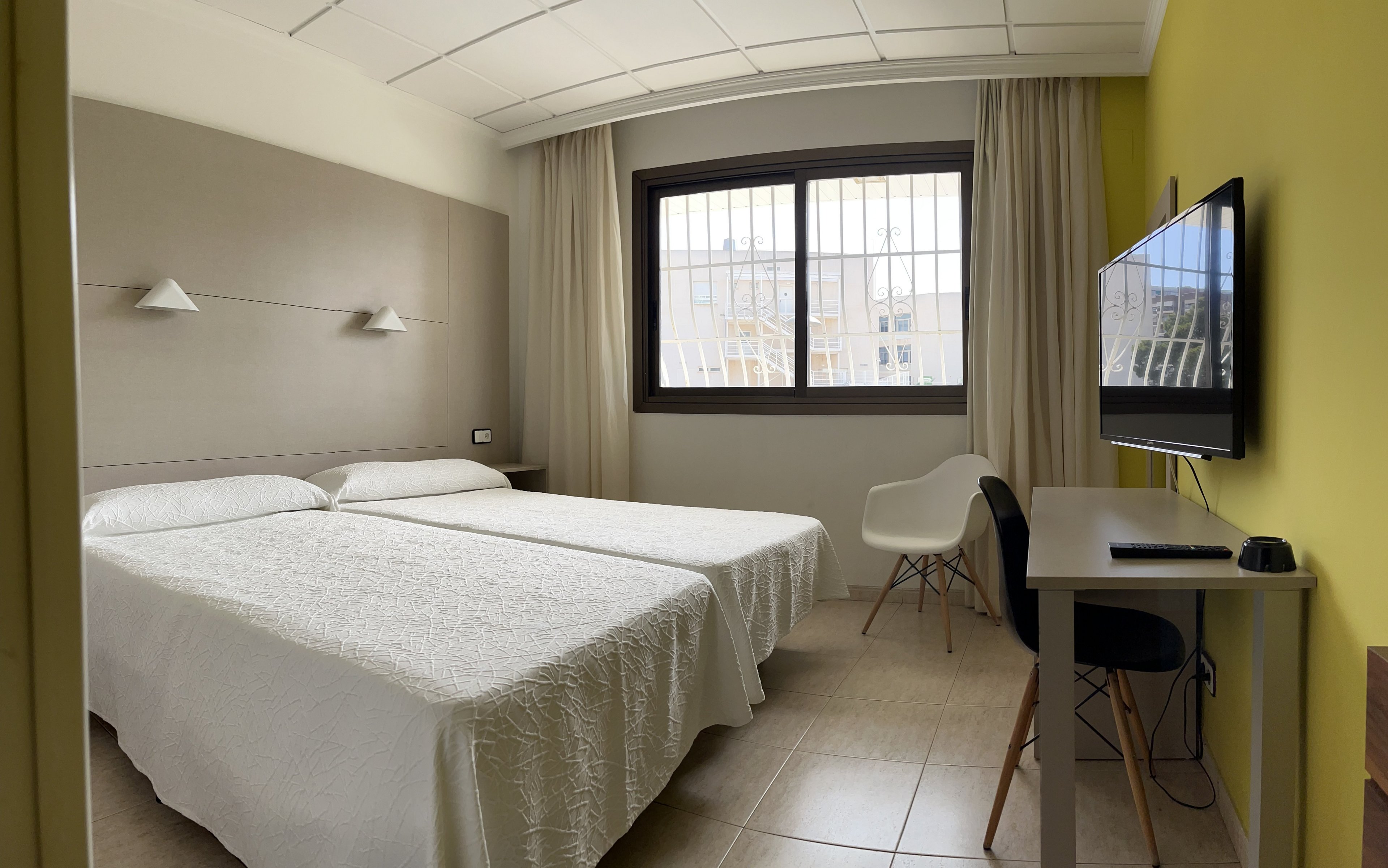 A compact, modern hotel room features two neatly made twin beds with white bedding against a light-colored headboard wall. A large window with bars and beige curtains lets in natural light, brightening the space. On the right side, a flat-screen television is mounted above a simple desk with a black chair and a small lamp. A white accent chair with wooden legs sits near the window, and the room is finished with tiled flooring and a yellow accent wall.