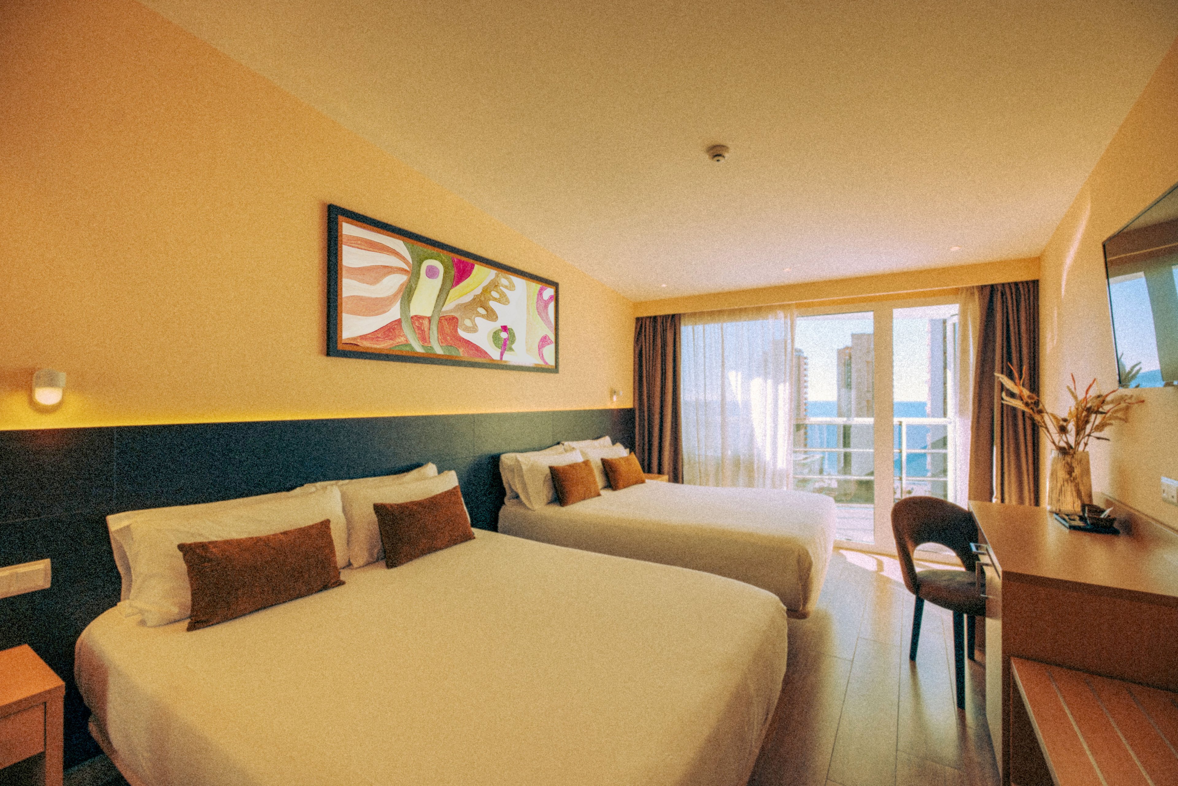 A modern hotel room features two large neatly made beds with white linens and brown accent pillows. Warm yellow walls and soft lighting create a cozy, inviting atmosphere. A desk with a chair and decorative vase sits opposite the beds, while a colorful abstract painting hangs above the headboard. Large glass doors open to a balcony with views of buildings and the sea in the distance.