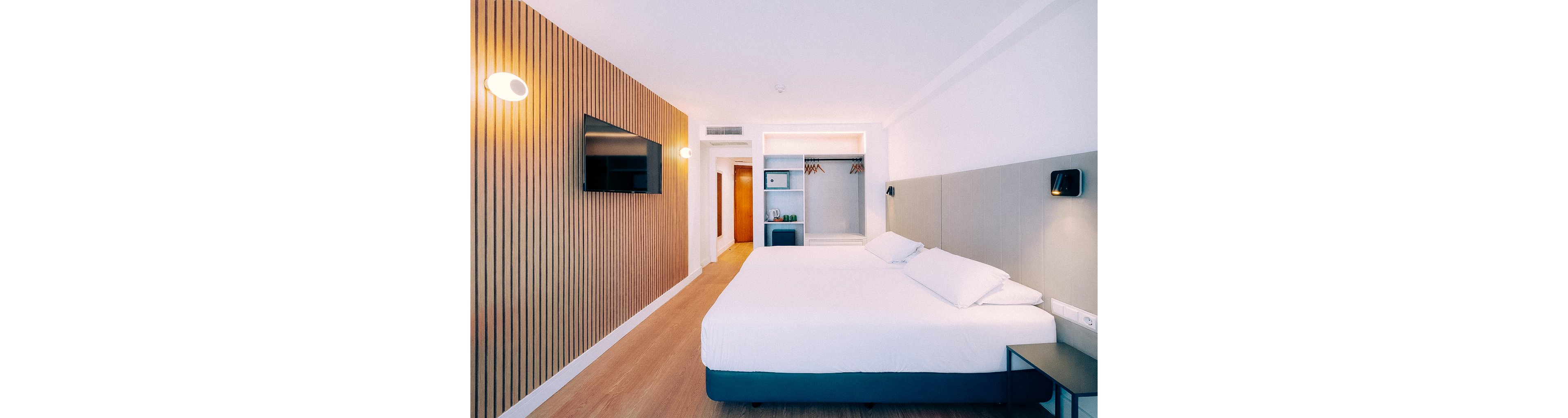Bright contemporary hotel room featuring two neatly made twin beds with white linens. The space includes a wooden accent wall with a mounted flat-screen television and minimalist bedside lighting. An open wardrobe, small refrigerator, and shelving unit are visible near the entrance door. The overall design is clean and modern, with light wood flooring and neutral color tones creating a calm atmosphere.