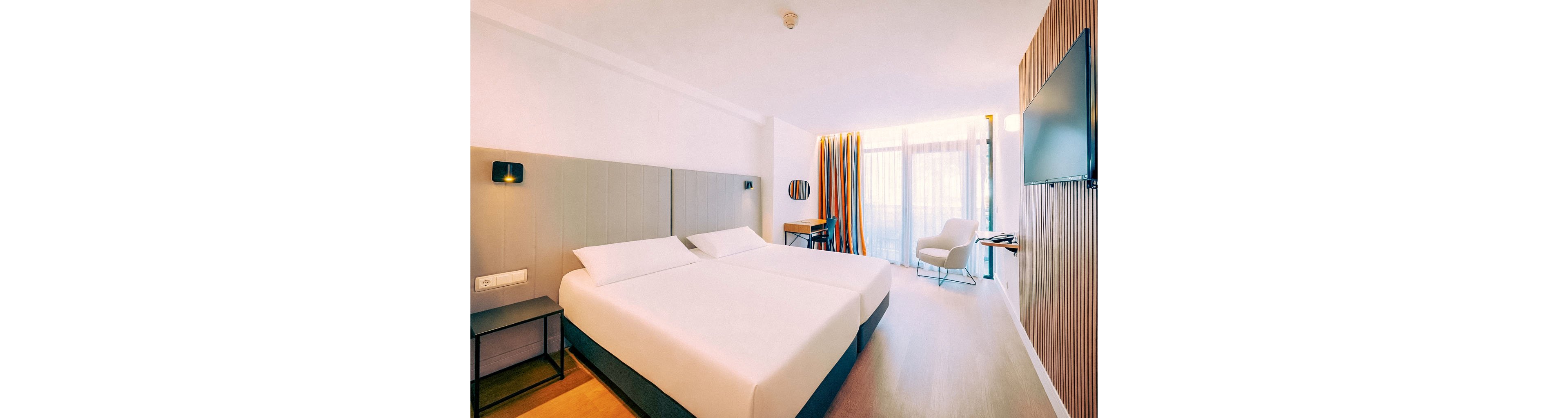 A bright, modern hotel room features two neatly made twin beds with a shared headboard. The space includes a small bedside table, a wall-mounted television, and a work desk with a chair near the window. Floor-to-ceiling windows with colorful striped curtains allow natural light to fill the room. A white armchair and minimalist decor create a clean, contemporary atmosphere.
