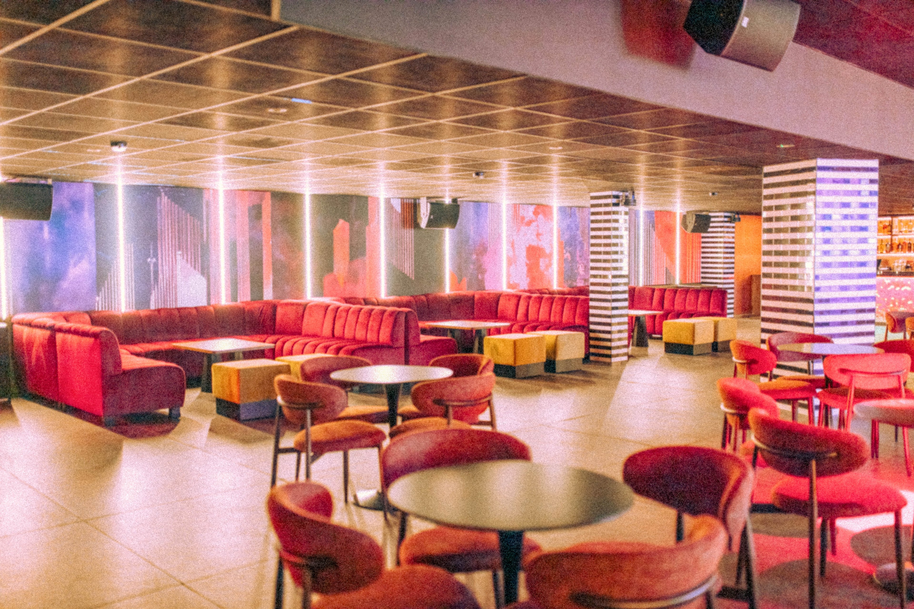 A contemporary lounge bar interior features red upholstered chairs and curved sofas arranged around small round tables. The space is empty, highlighting the layout of seating areas and open floor. Vertical light columns and striped pillars add a stylish, modern touch to the room. Warm ambient lighting creates a cozy, inviting nightlife atmosphere.