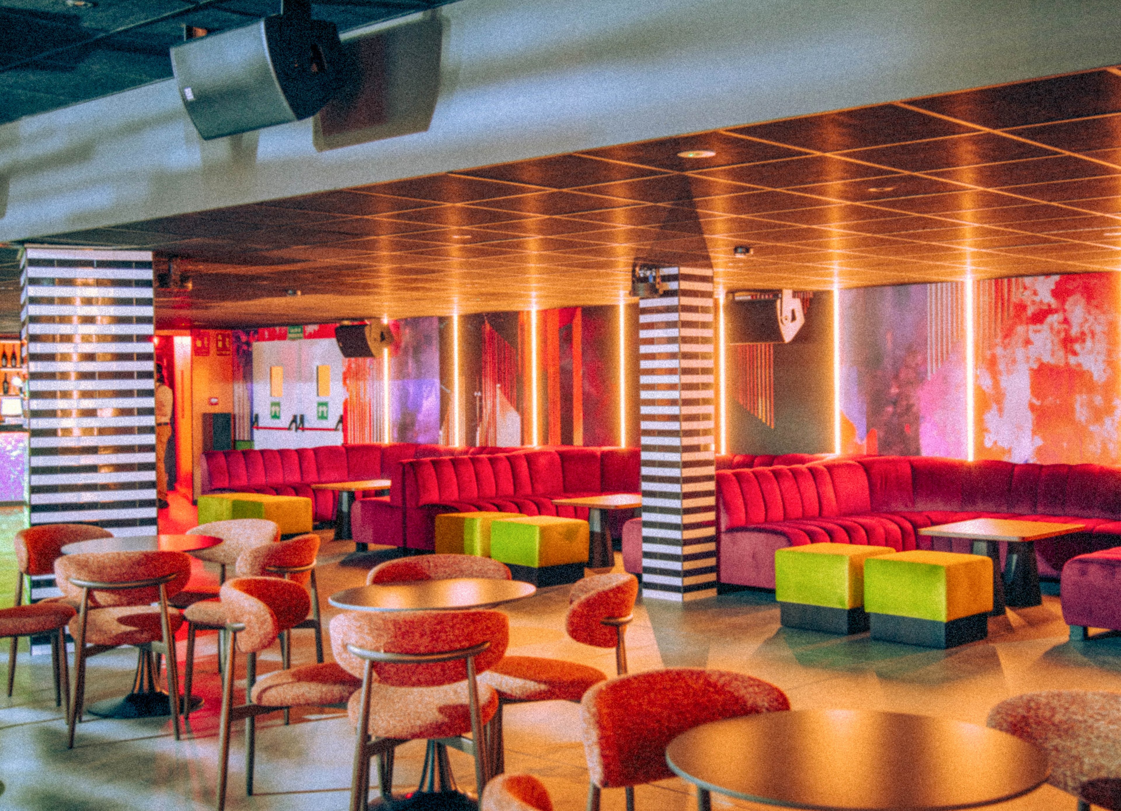A vibrant lounge bar interior features rows of red upholstered banquettes and round tables with cushioned chairs. The space is empty, highlighting the geometric layout of seating and the striped columns that divide the room. Warm ambient lighting in orange and pink tones reflects off the walls, creating a colorful, nightlife atmosphere. Neon-like hues and bold color blocks on the stools add to the contemporary, trendy club feel.