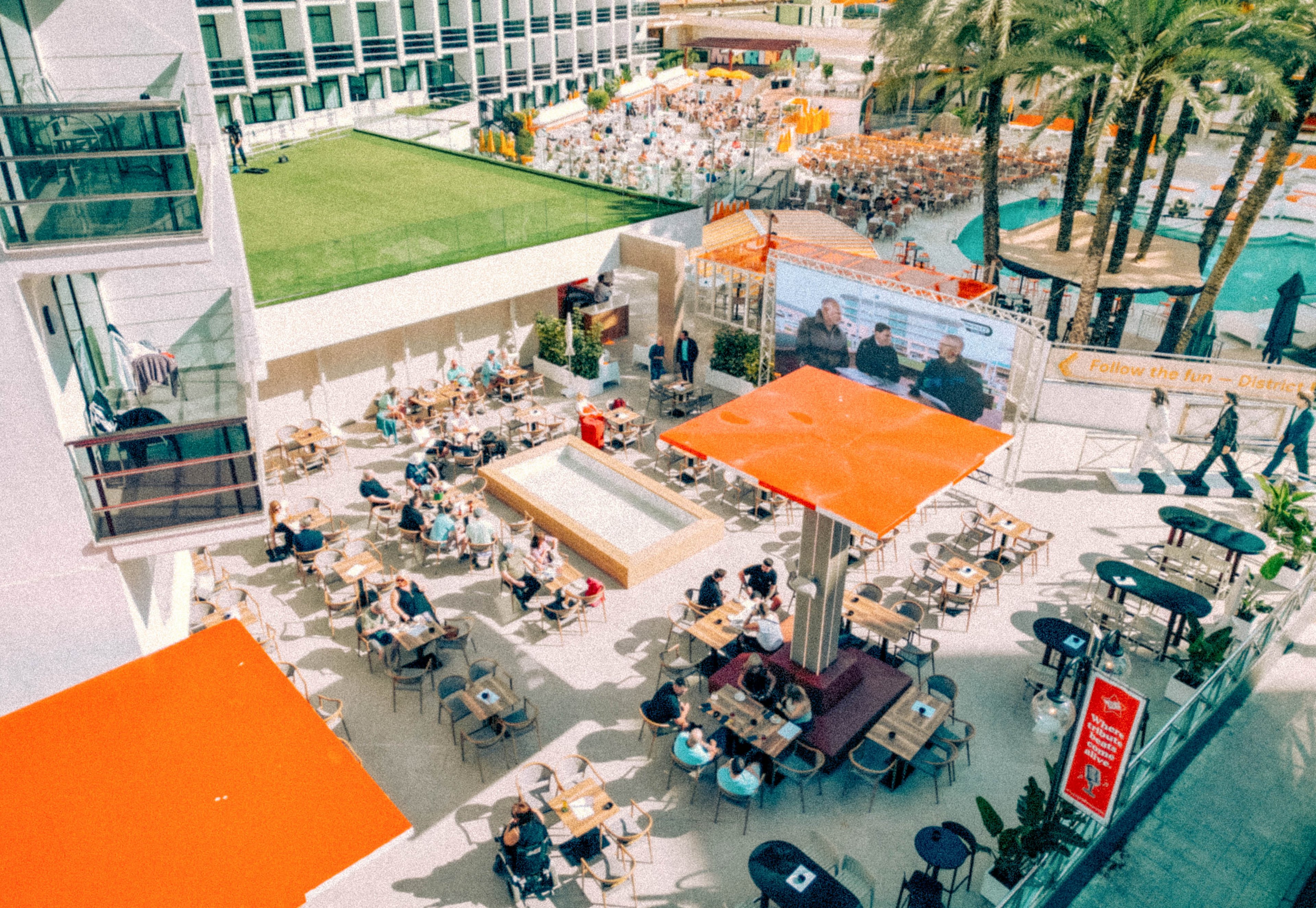 An elevated view shows a busy outdoor hotel terrace filled with people sitting at small tables. Bright orange umbrellas and sunshades provide color contrast against the light stone flooring. In the background, more guests relax near a swimming pool and palm trees, with a large screen displaying an event or broadcast. The scene suggests a lively vacation atmosphere in a warm, sunny resort setting.