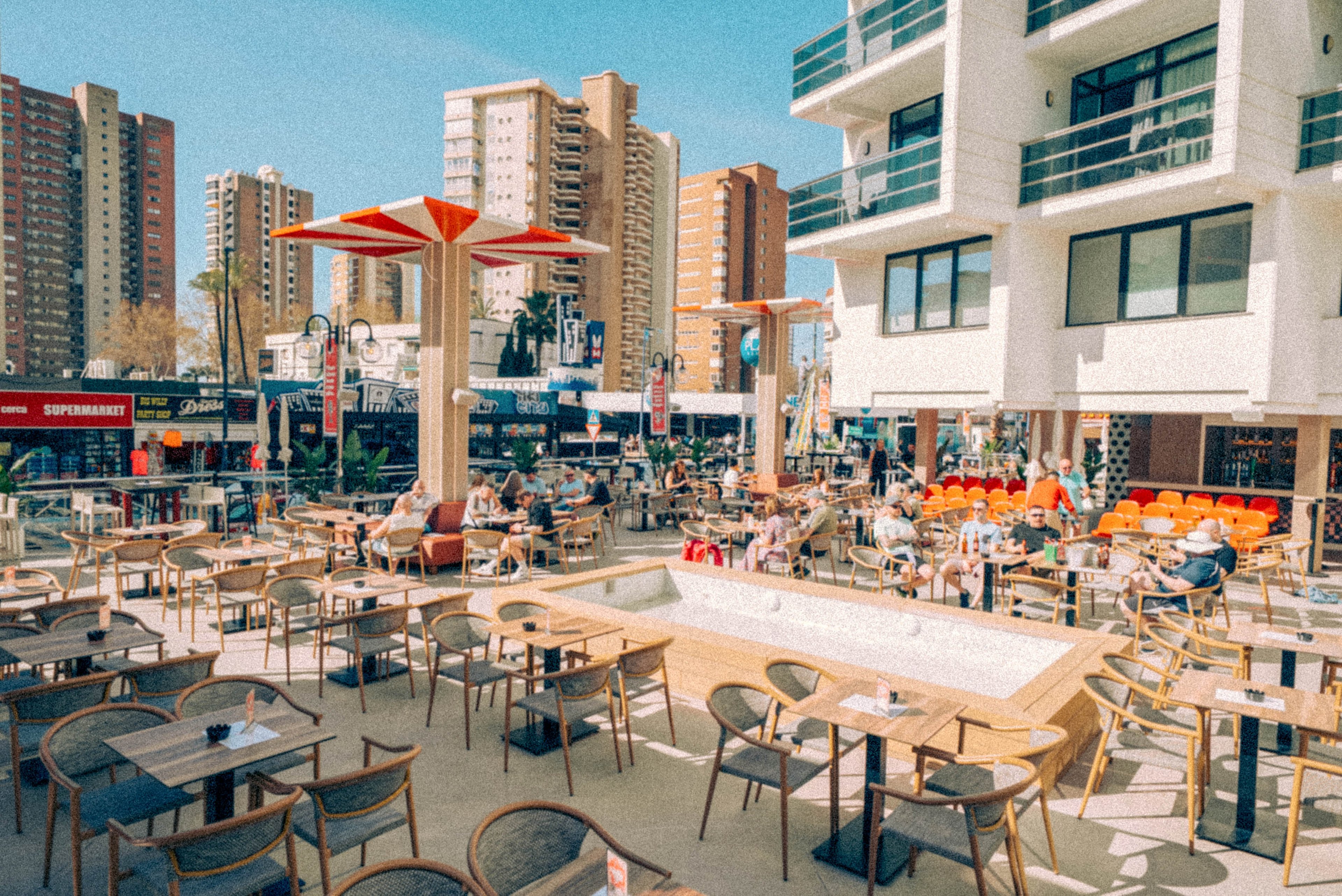 An expansive outdoor cafe terrace is filled with tables and chairs under a bright blue sky. People sit and relax at the cafe while tall modern apartment buildings rise in the background. Distinctive orange and white shade structures stand over the seating area, adding a contemporary touch. The overall scene suggests a lively urban square with a relaxed, holiday atmosphere.