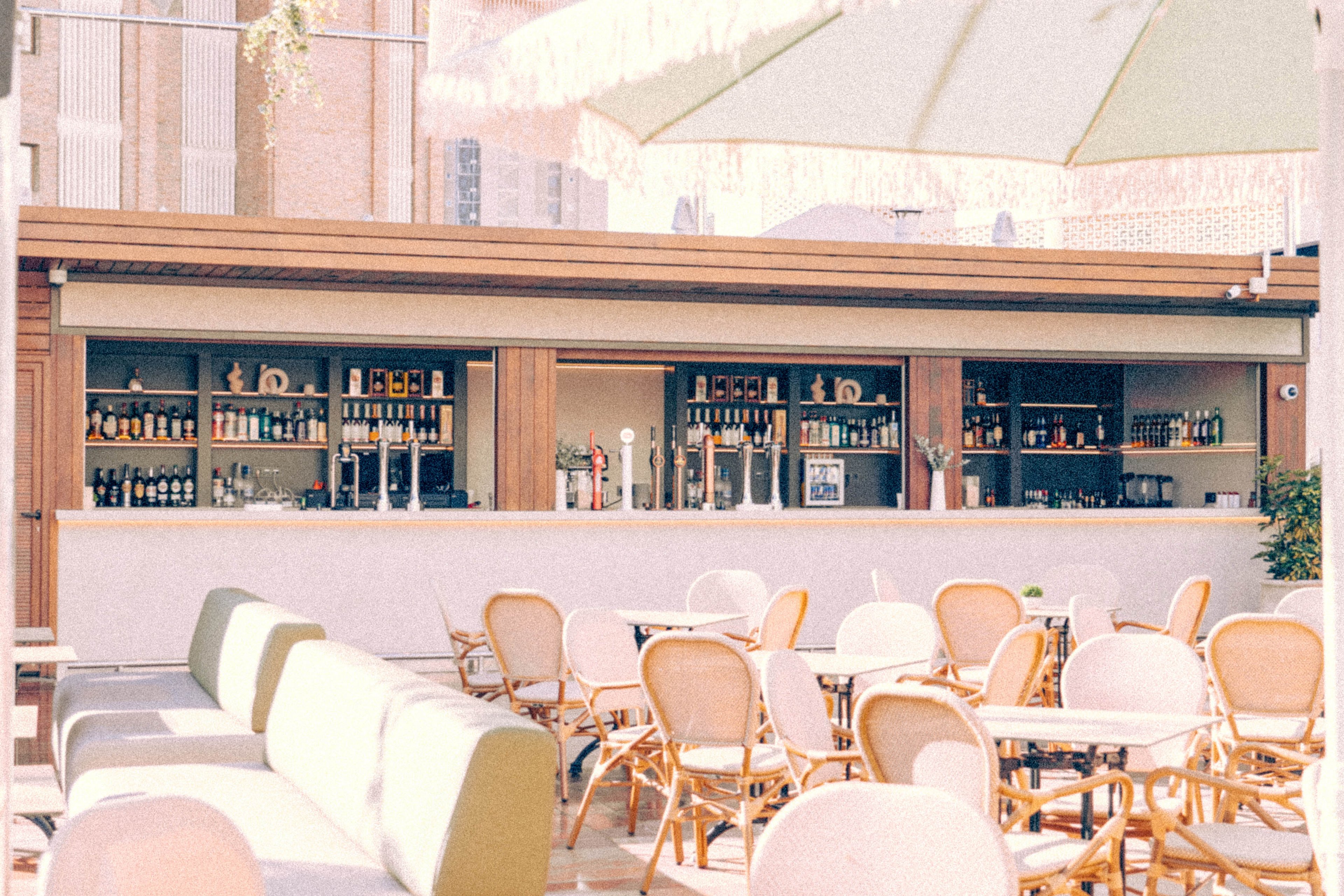 An open-air rooftop bar features a long counter lined with bottles and drink dispensers. In the foreground, light-colored chairs and tables are arranged for guests on a sunlit terrace. A cushioned bench and large patio umbrella add comfort and shade to the seating area. Tall city buildings rise in the background, creating an urban atmosphere. The overall scene feels relaxed, bright, and inviting for daytime socializing.