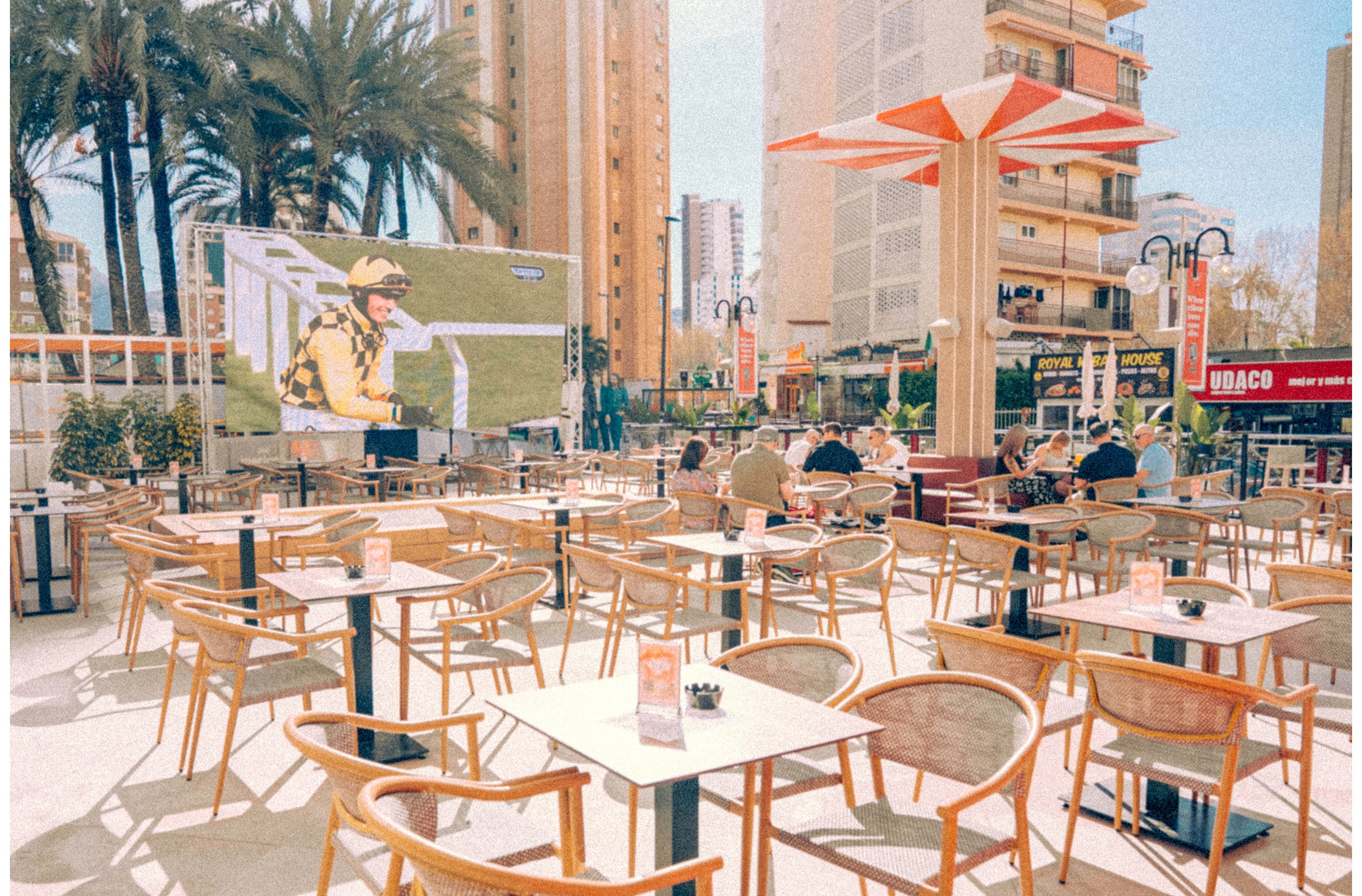 An open-air café terrace features many empty wooden chairs and tables arranged in neat rows. A few people sit near the back, facing a giant outdoor screen showing a sports broadcast with a person in yellow attire. Tall palm trees and high-rise buildings surround the seating area, creating an urban holiday atmosphere. Warm sunlight and soft pastel tones give the scene a relaxed, summery mood.