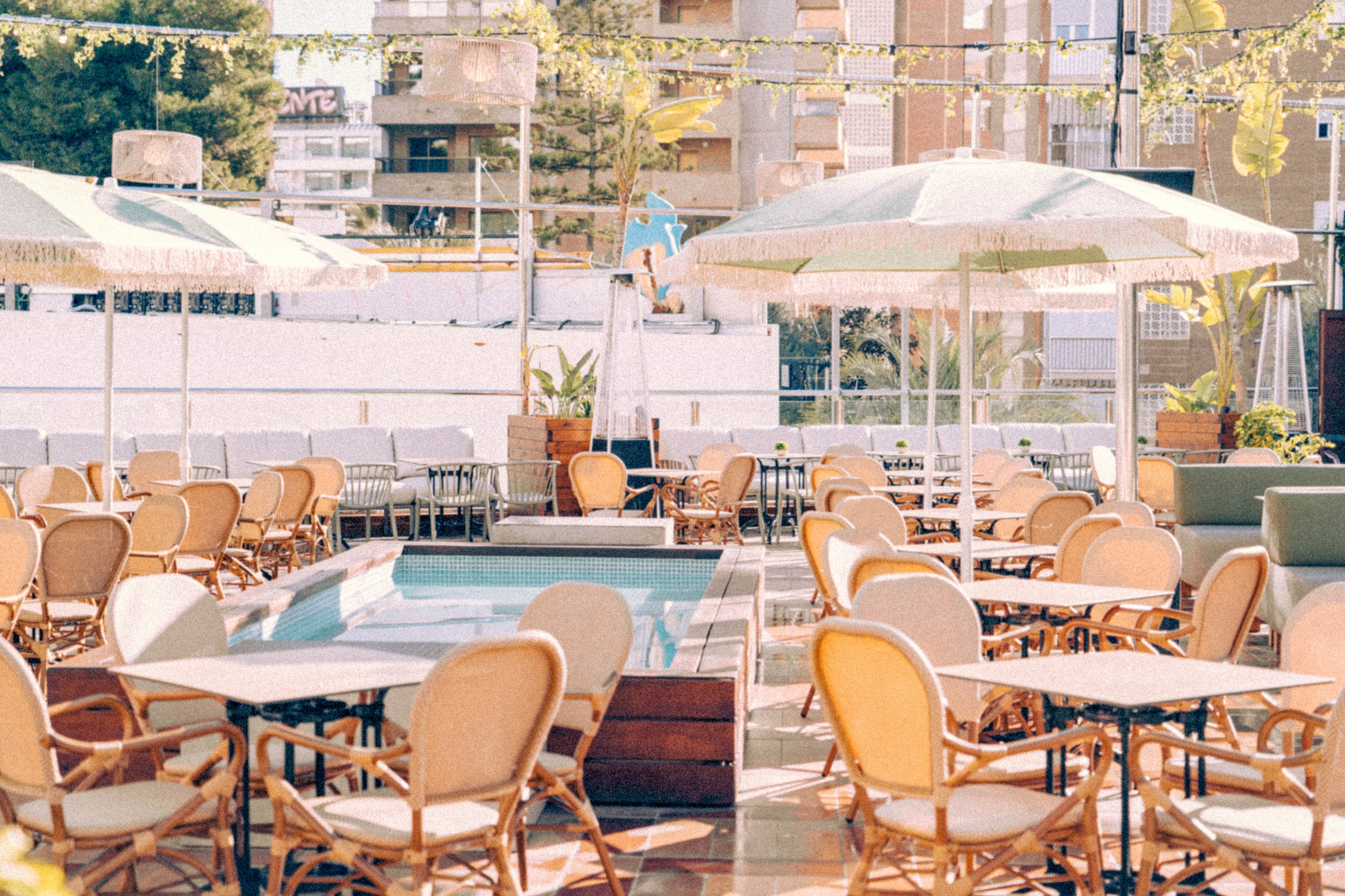 An outdoor café terrace is filled with empty tables and wicker chairs under large green-and-white umbrellas. The scene is bathed in warm sunlight, creating soft reflections on the polished floor. In the background, urban apartment buildings and greenery frame the relaxed setting. The overall atmosphere suggests a quiet, inviting place to sit and enjoy a drink or meal.