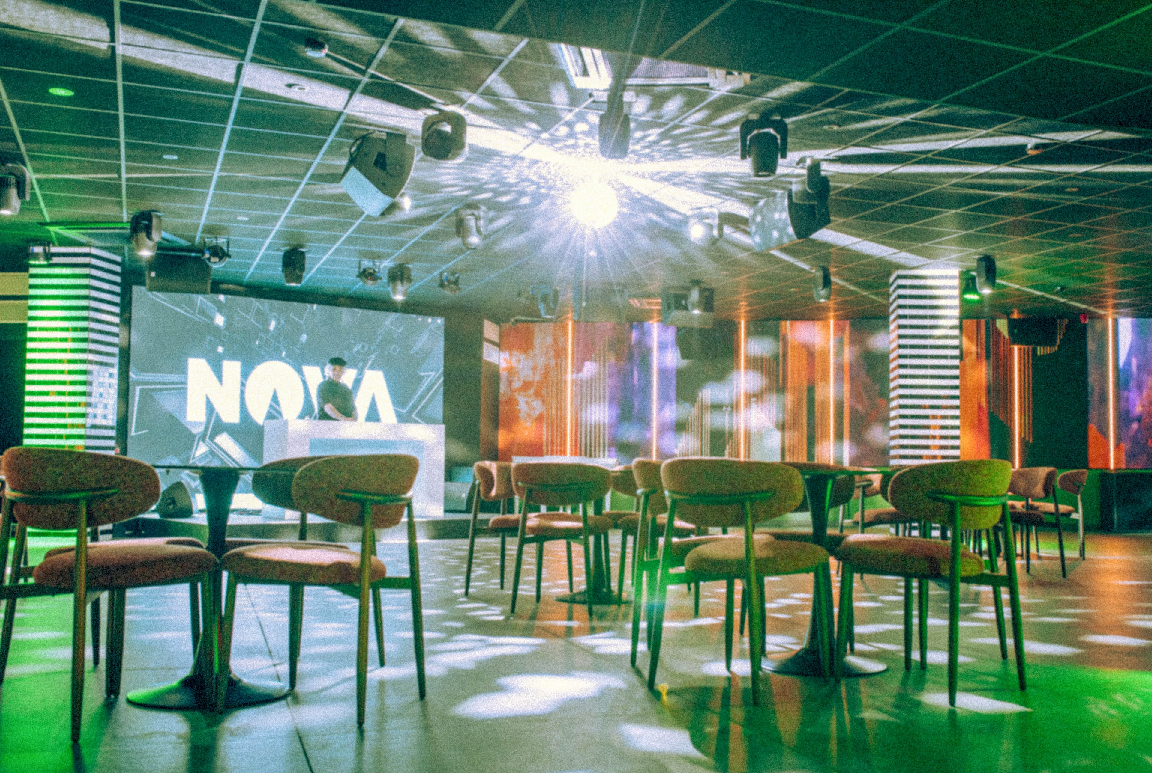 A modern nightclub interior features a DJ booth in front of a large screen displaying the word NOVA. Rows of empty chairs and small tables are arranged across the dance floor, suggesting the venue is ready for guests. Colorful LED panels and spotlights cast green, orange, and white hues throughout the space. A bright light near the ceiling creates starburst reflections, enhancing the energetic, futuristic atmosphere.