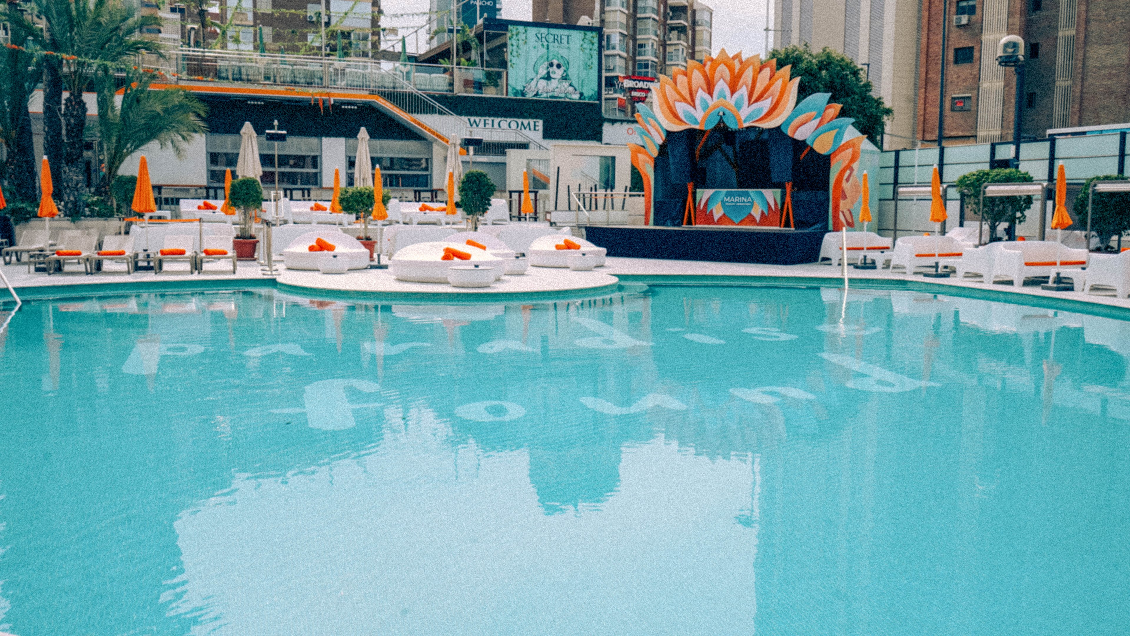 A large turquoise swimming pool is surrounded by white lounge beds and bright orange umbrellas. At the far end, a decorative stage with bold orange and blue floral shapes creates a focal point. The setting appears to be an urban rooftop or terrace, with tall buildings and billboards visible in the background. The scene feels calm and stylish, suggesting a resort or day club atmosphere.
