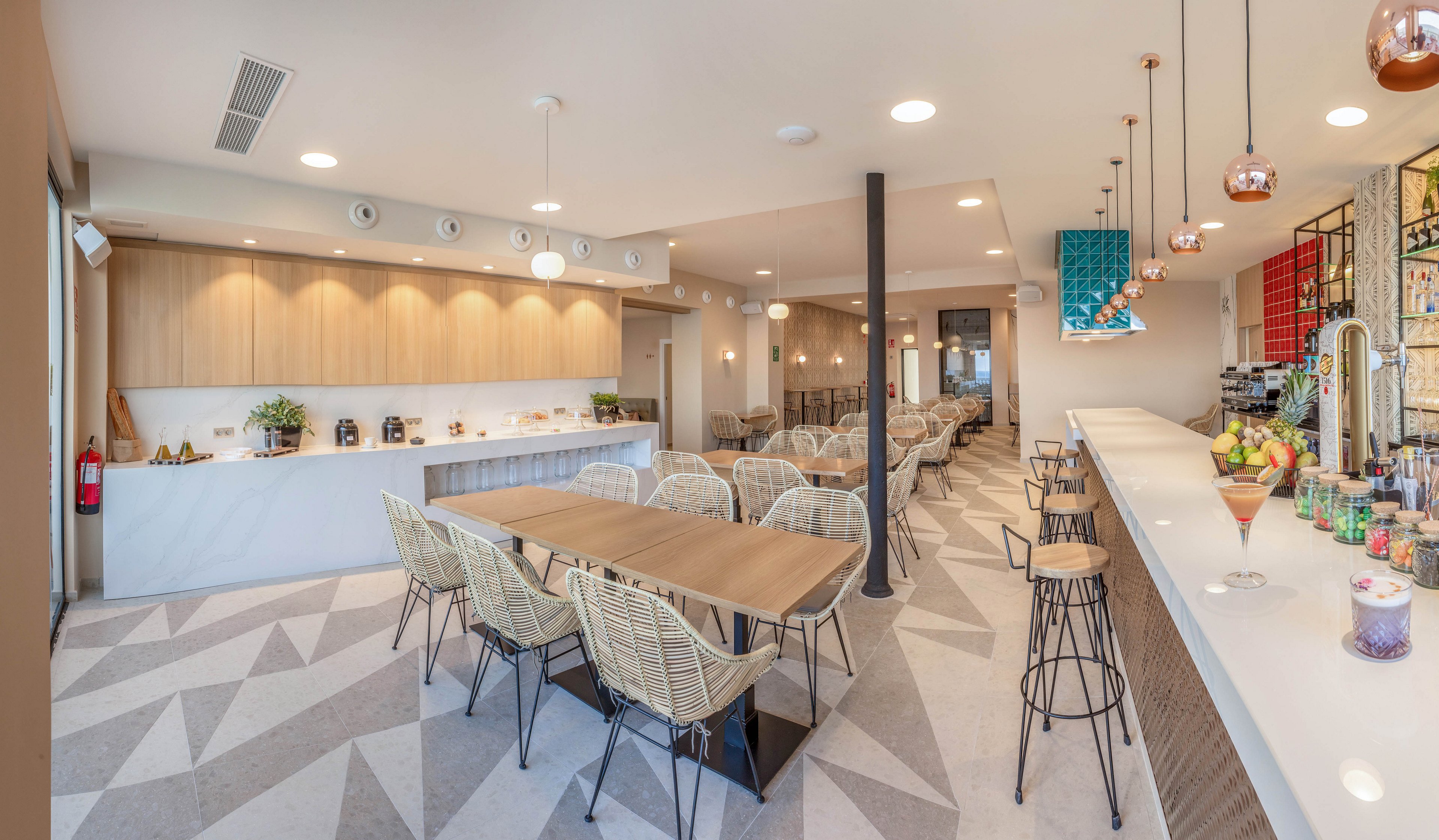 Bright contemporary cafe interior featuring a long bar counter and communal tables. Woven chairs and minimalist wooden cabinetry create a warm, inviting atmosphere. Geometric patterned flooring and pendant lights add visual interest to the open space. The bar area displays drinks and fruit, suggesting a casual dining or coffee shop setting.
