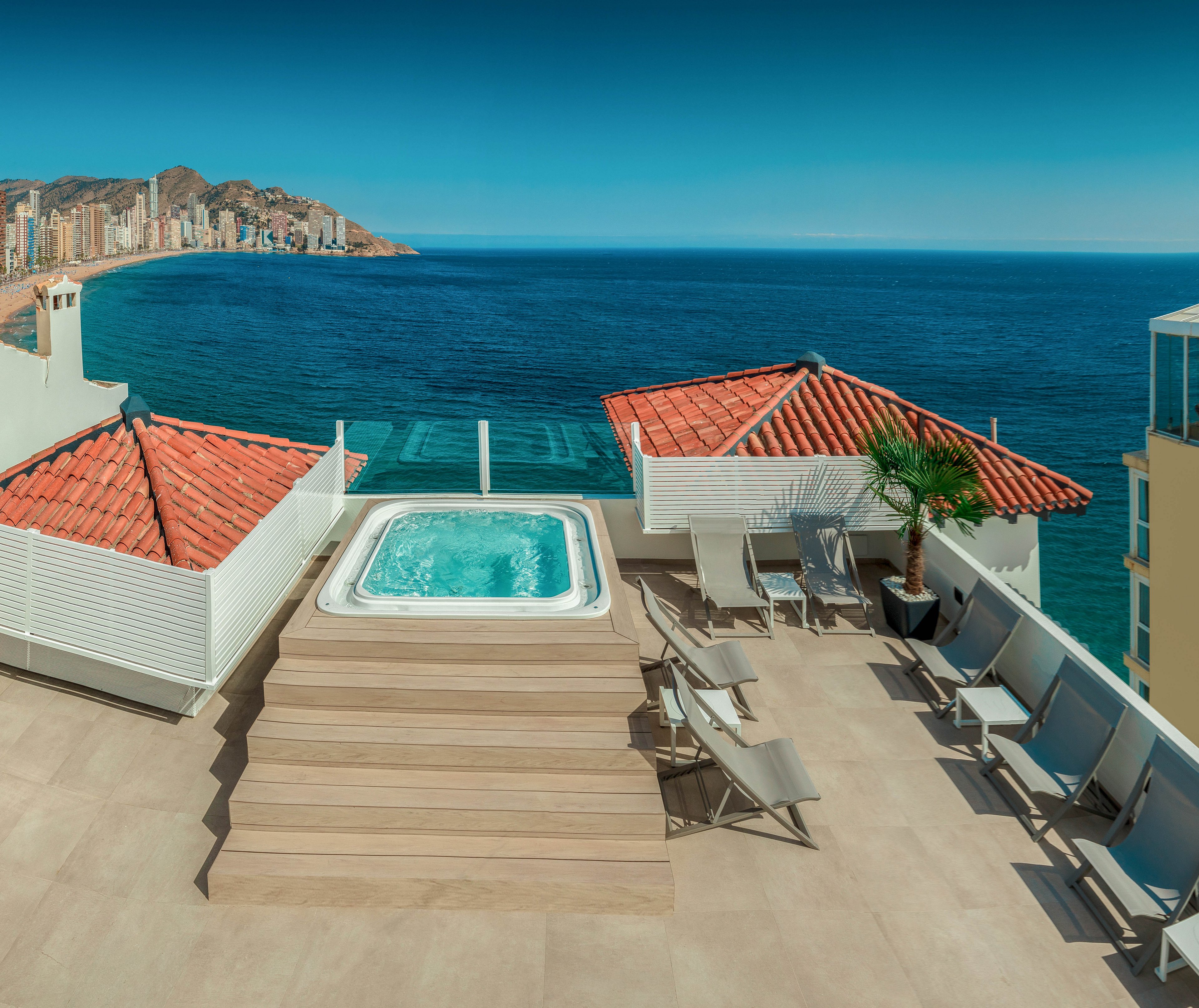 A modern rooftop terrace features a rectangular hot tub centered on a raised wooden platform. Surrounding the spa are lounge chairs and seating areas, framed by white walls and red-tiled roofs. A glass railing opens to a panoramic view of the deep blue sea and a distant coastal city skyline. The clear sky and bright sunlight create a serene, luxurious vacation atmosphere.
