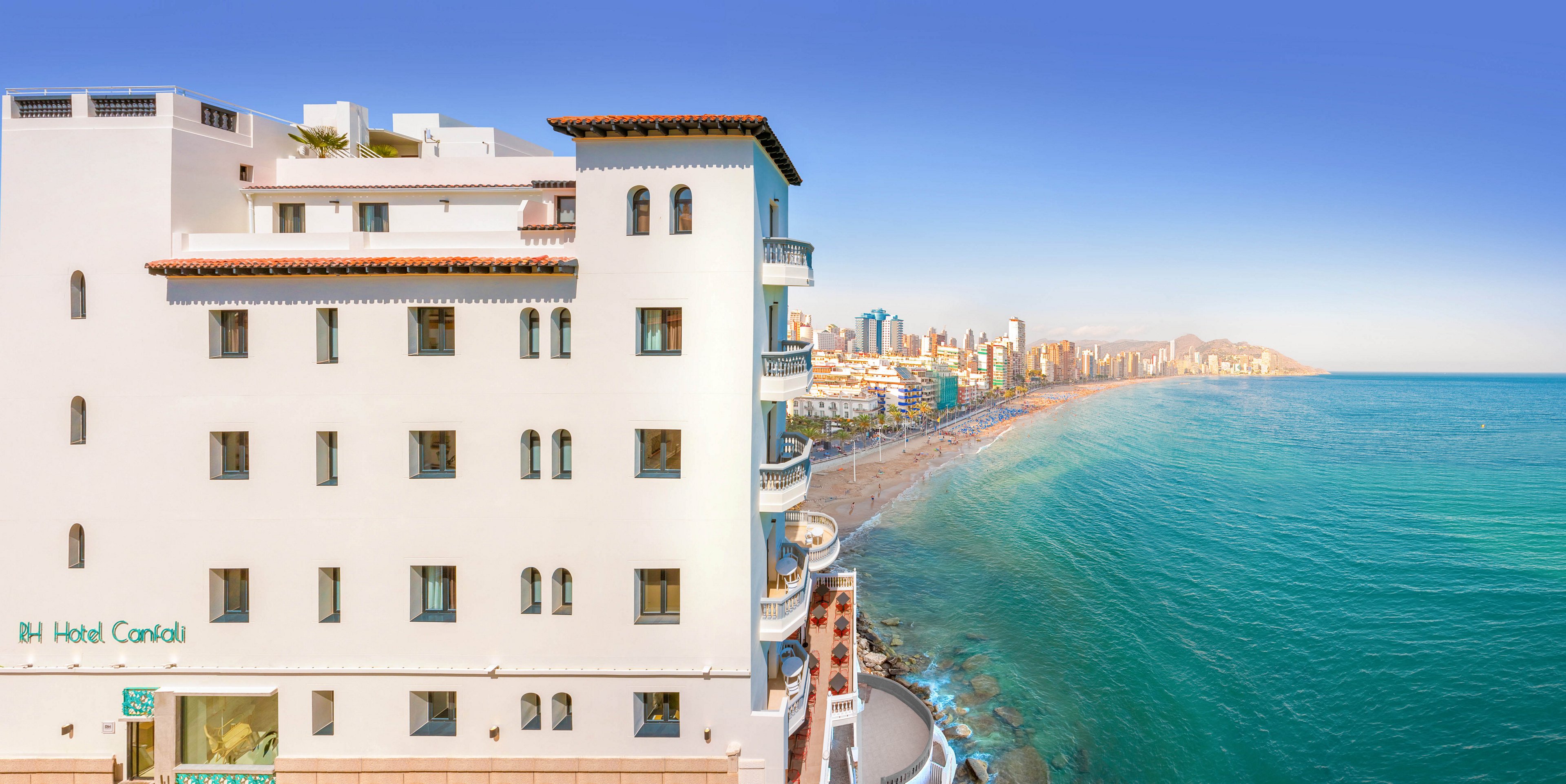 A white multi-story seaside hotel stands prominently on the left side of the image. The building faces a long sandy beach that curves along a turquoise sea. In the distance, a dense skyline of modern high-rise buildings lines the coast under a clear blue sky. The scene suggests a warm, sunny day in a popular coastal city resort.