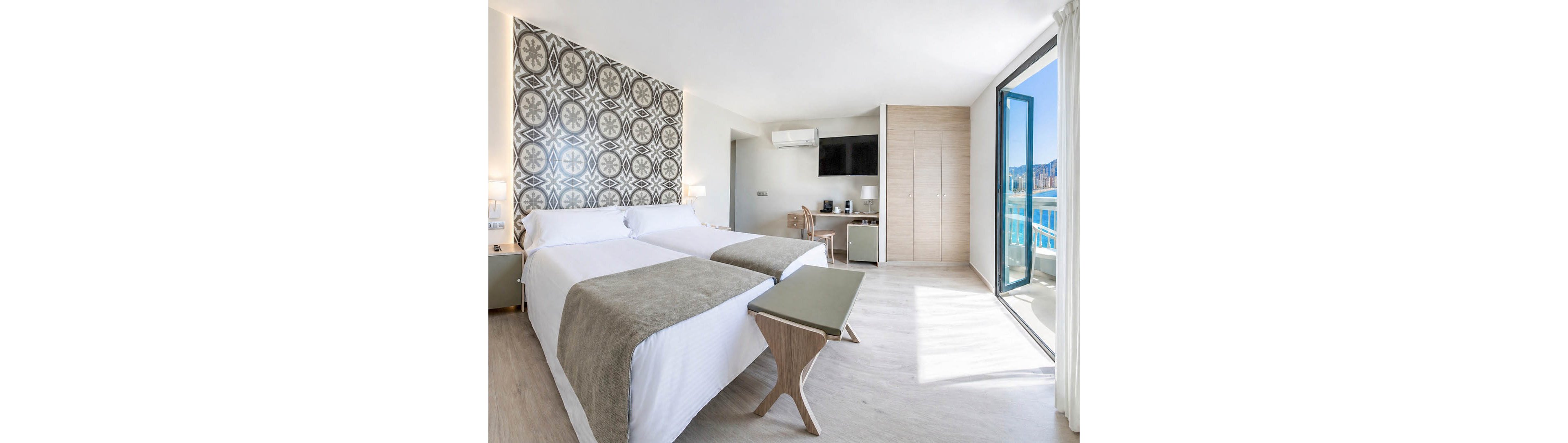Spacious contemporary hotel room featuring two neatly made twin beds with light gray throws. The space is bright and airy, with large windows and a glass door opening to a sunny balcony and coastal view. A patterned accent wall, minimalist wooden furniture, and neutral tones create a clean, modern aesthetic. A small desk area with a television and compact refrigerator completes the functional layout.