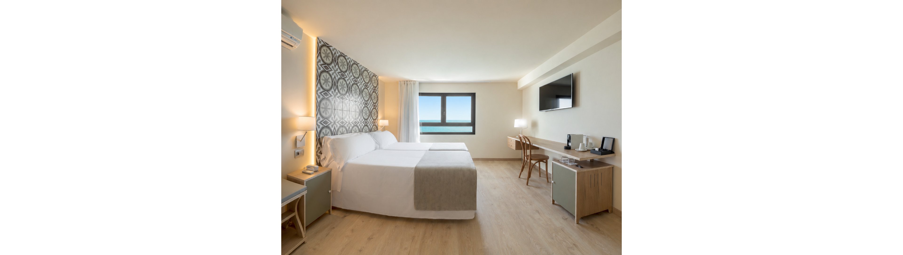 A bright, modern hotel room features two neatly made beds with light gray runners and a decorative patterned headboard. A large window in the center of the far wall offers a clear view of the ocean and blue sky. On the right side, a wooden desk with a chair holds a flat-screen TV, lamps, and a coffee and tea station. Soft neutral tones, clean lines, and ample natural light create a calm, minimalist atmosphere.