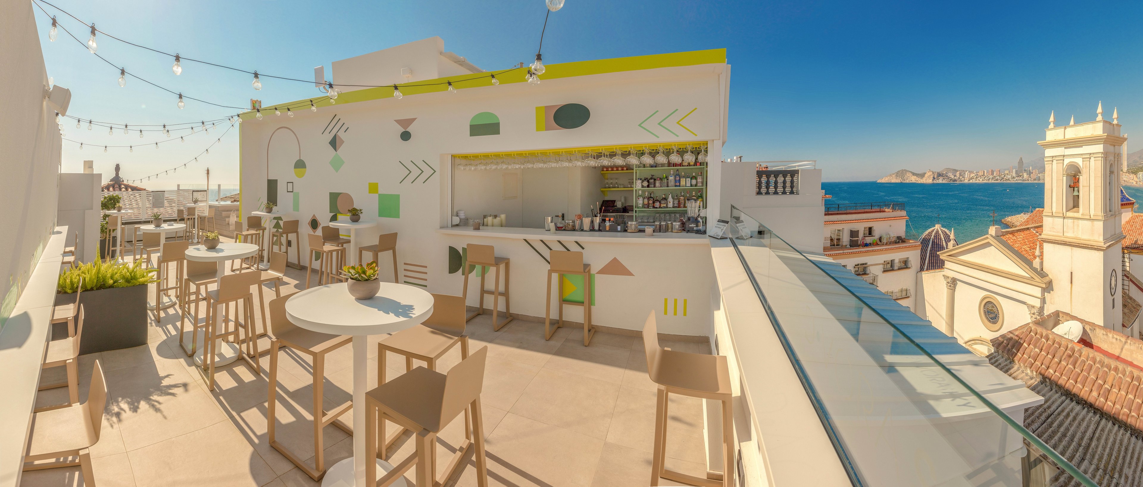 Open-air rooftop bar with modern white furniture sits above a coastal town under a clear blue sky. High stools and round tables line the terrace, facing a compact bar counter stocked with bottles and glassware. Decorative geometric shapes and pastel accents brighten the minimalist walls, while string lights hang overhead for evening ambiance. In the background, historic buildings and a church tower rise above red-tiled roofs, with the sea and distant shoreline visible beyond.