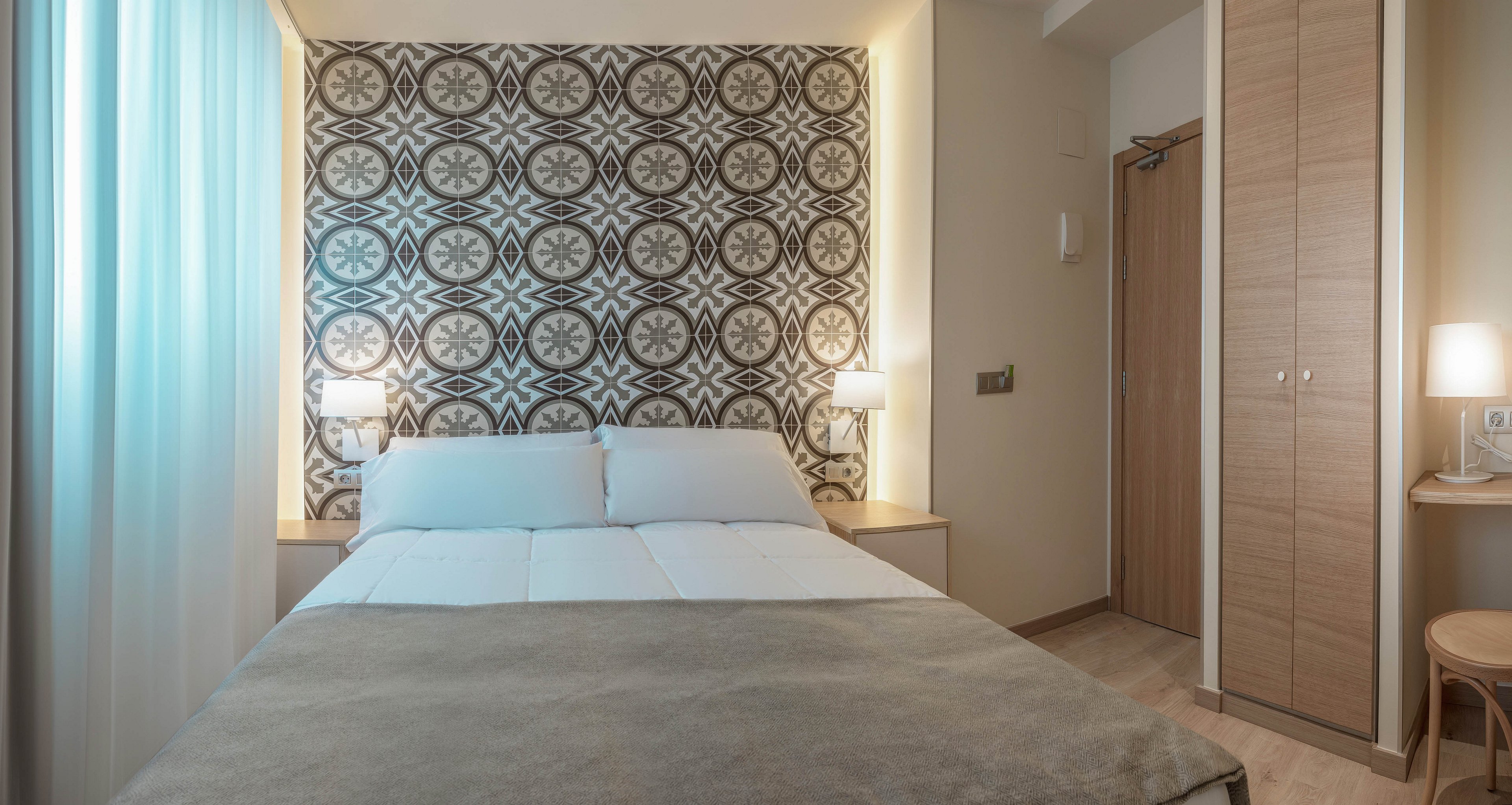 A neatly made double bed sits centered in a compact, modern bedroom. The headboard wall features a bold geometric pattern with circular floral motifs, framed by recessed lighting. Soft bedside lamps and sheer curtains create a warm, inviting glow. A tall wooden wardrobe and light neutral tones emphasize a clean, minimalist design.