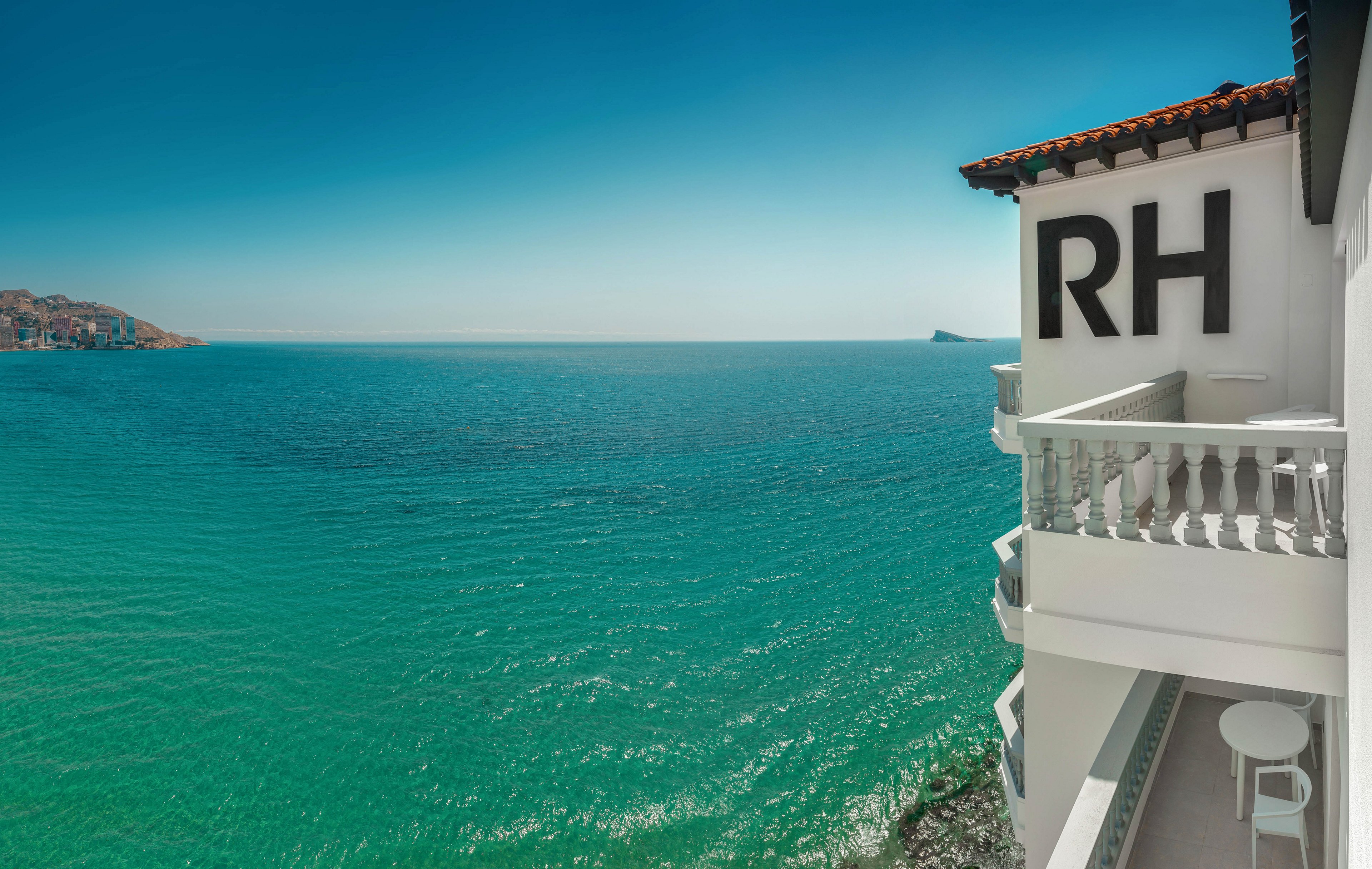 A white seaside building with the large letters RH on its facade overlooks a calm turquoise sea. The view from the balcony shows clear water meeting a distant coastline under a bright blue sky. Classic white railings and a small round table emphasize the relaxed coastal setting. The scene conveys a tranquil, sunny holiday atmosphere on the waterfront.