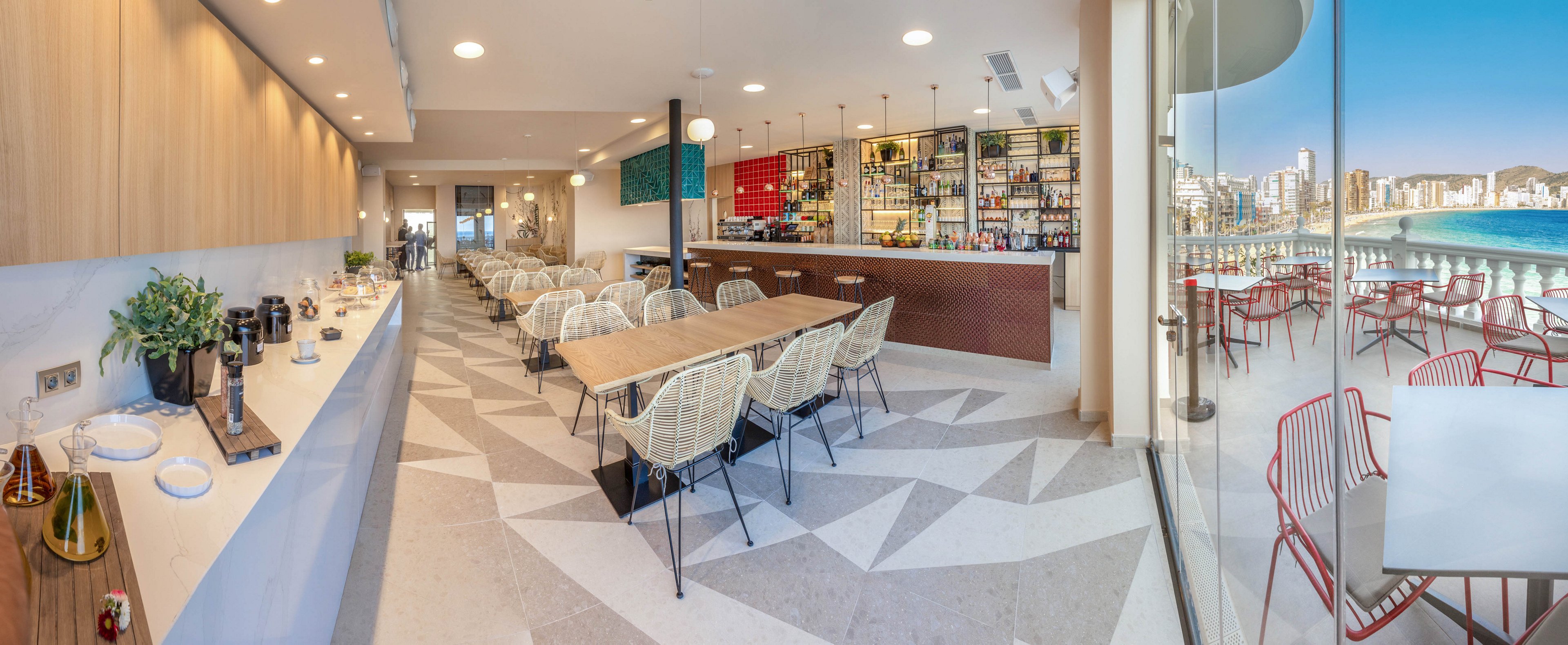 Contemporary restaurant interior featuring a long wooden bar and neatly arranged dining tables with woven chairs. The space is bright and airy, with geometric patterned flooring and warm wood paneling along one wall. A self-service counter with coffee equipment and plants runs parallel to the seating area. Large glass windows open to an outdoor terrace with red chairs and a scenic view of the beach and city skyline.