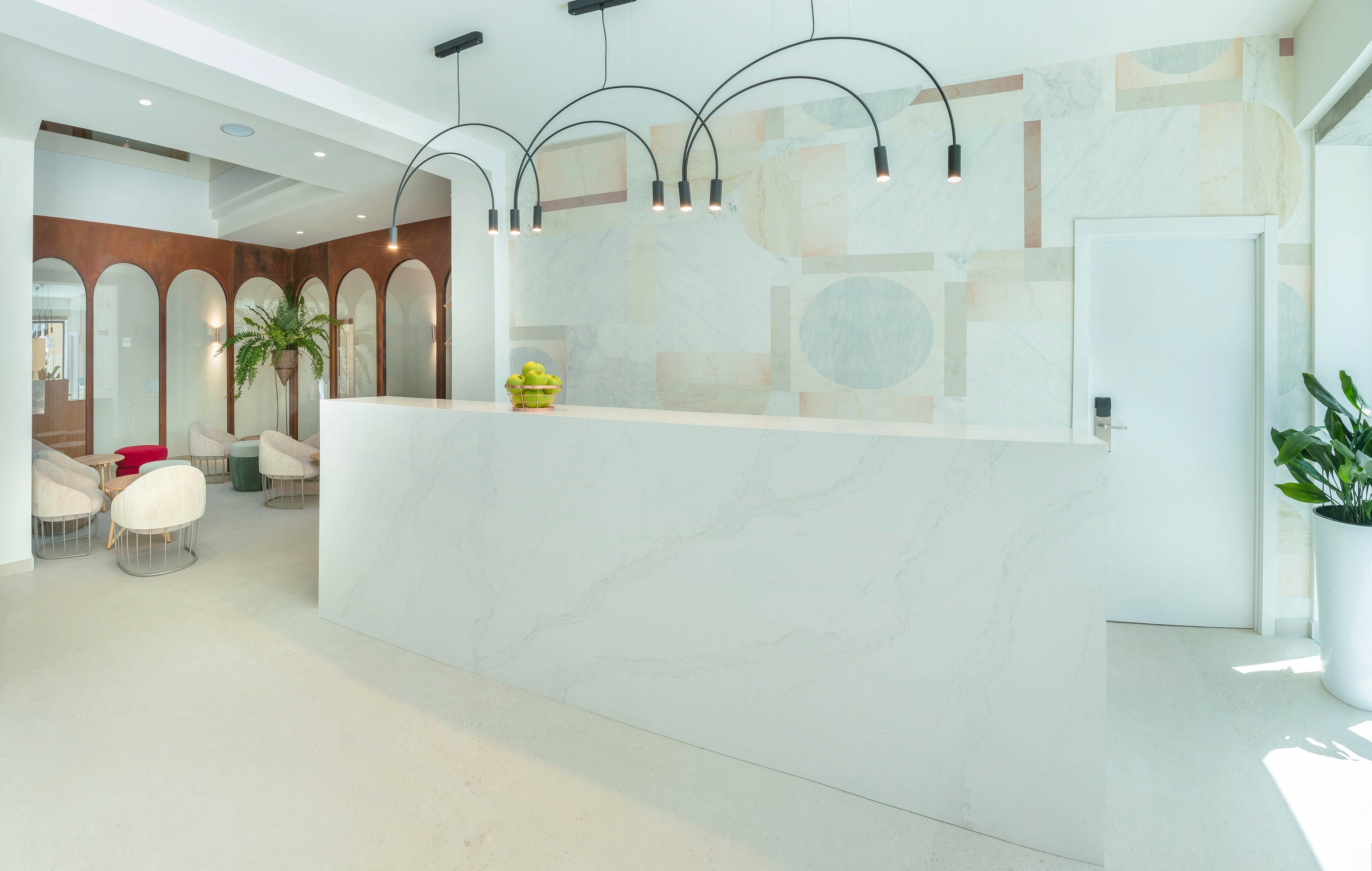 A bright, modern reception area features a large white marble-style counter as the central focus. Curved black pendant lights hang above, illuminating the clean, minimalist space. In the background, arched wall openings reveal a cozy seating area with upholstered chairs and a tall indoor plant. Soft neutral tones, geometric wall patterns, and natural light create an airy, contemporary atmosphere.