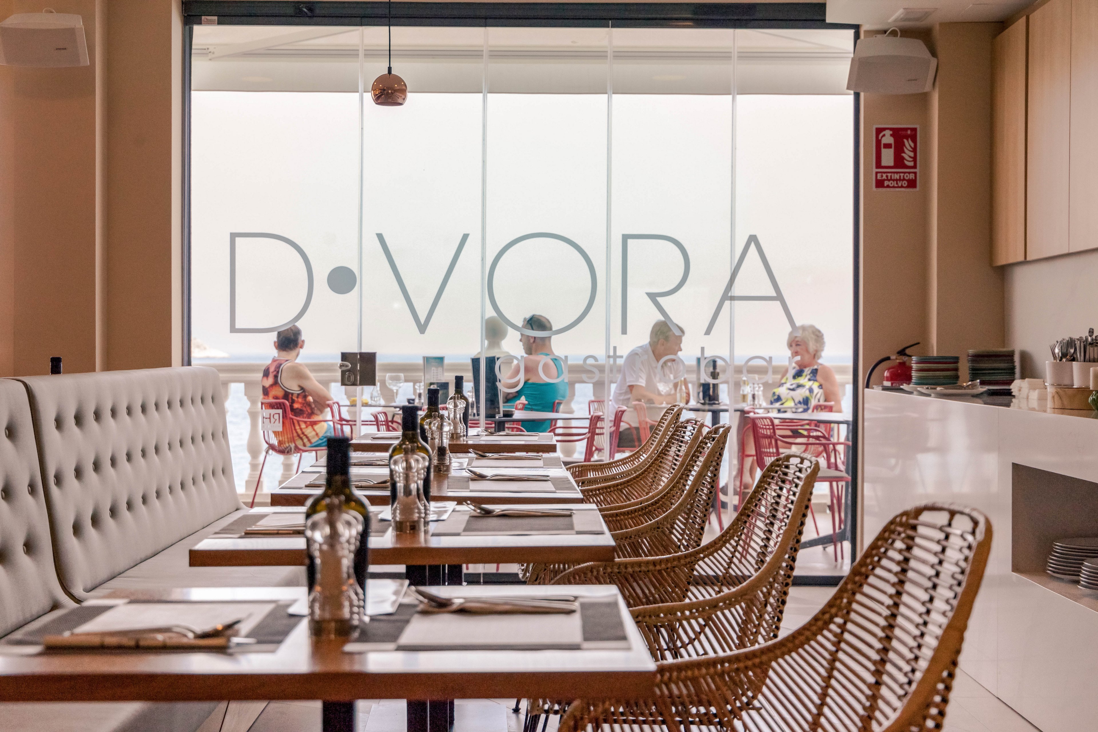 Contemporary restaurant interior with neatly set wooden tables and wicker chairs faces large floor-to-ceiling windows. The foreground shows place settings with cutlery, napkins, and bottles of oil or vinegar arranged in the center of each table. Through the glass, several people sit on the outdoor terrace, enjoying a bright seaside view. Soft neutral tones and natural materials create a relaxed, modern dining atmosphere. Large lettering on the window reads "D·VORA" above the terrace area.