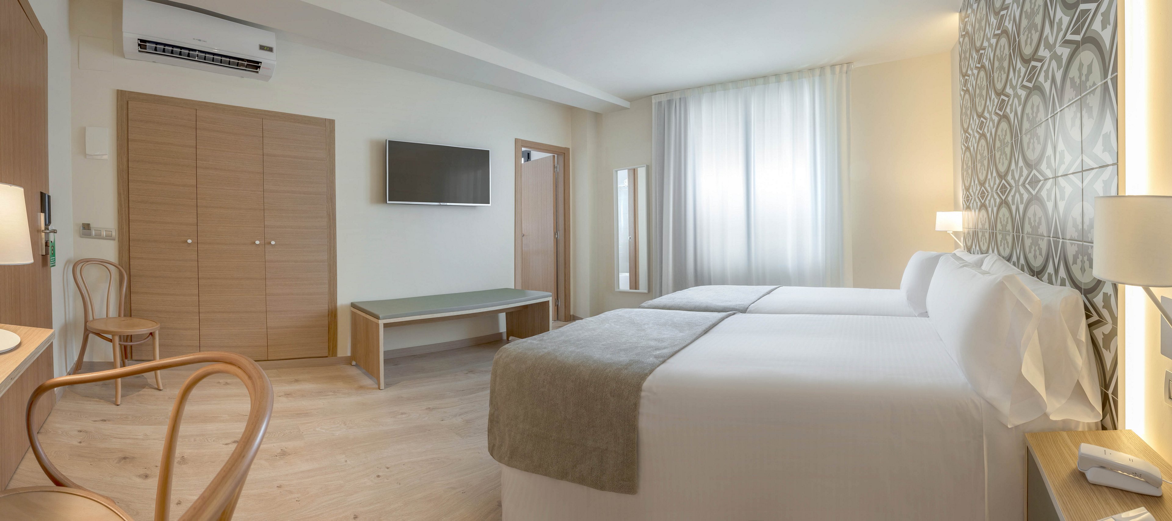 A contemporary hotel room features two neatly made twin beds with white linens and light gray throws. The space is bright and airy, with natural light streaming through sheer curtains covering a large window. A flat-screen television is mounted on the wall opposite the beds, above a minimalist bench. Light wood furniture, a patterned accent wall, and neutral tones create a calm, modern atmosphere.