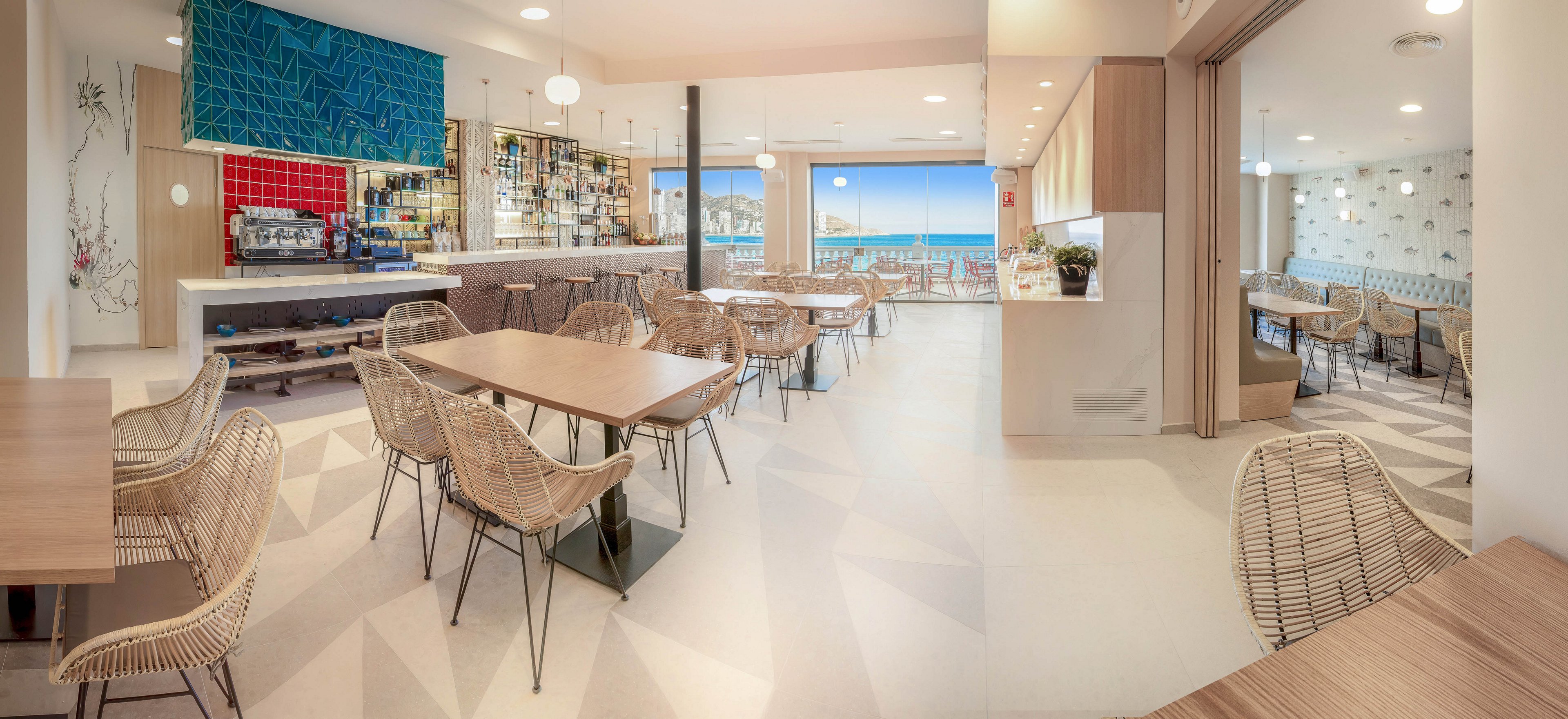 Spacious modern restaurant interior featuring light wooden tables and woven chairs. A bar area with colorful tiles and neatly arranged bottles stands to the left. Large windows open to a terrace with views of the ocean and distant coastline. Neutral tones, ample natural light, and clean lines create an airy, contemporary dining atmosphere. The space appears empty, ready to welcome guests.