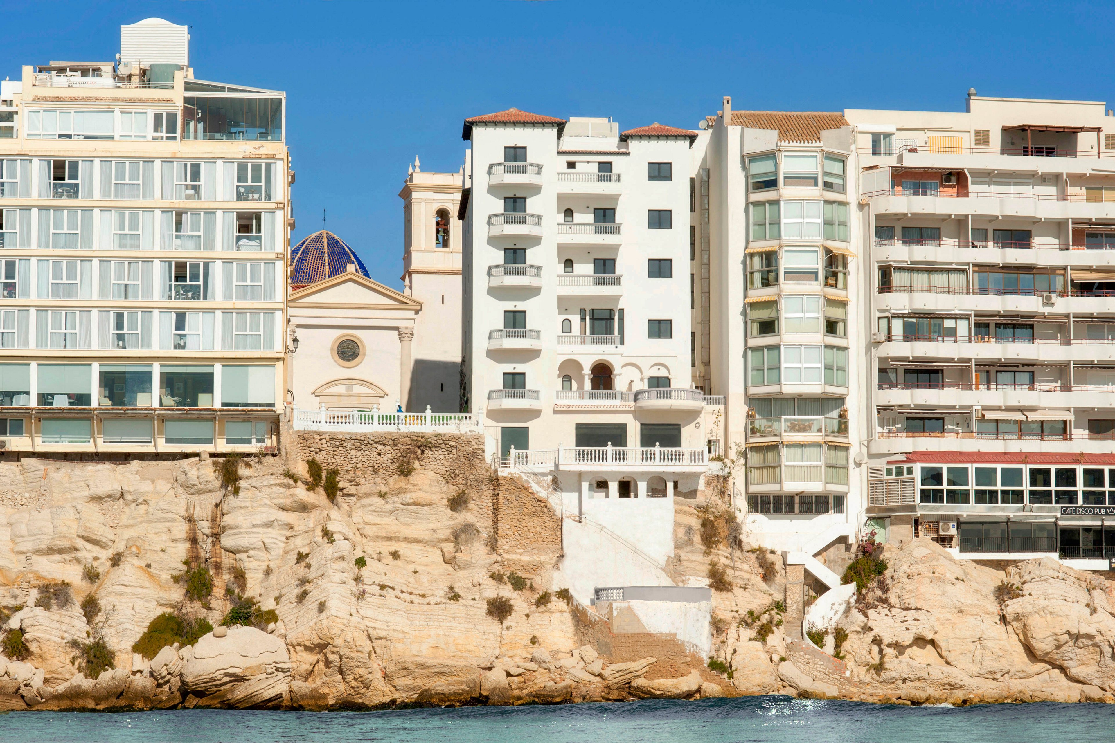A row of mid-rise apartment buildings stands atop a rocky seaside cliff. Among the modern structures, a small church with a dome roof and bell tower adds historic character. Sunlit facades and balconies overlook the blue water below. The scene suggests a Mediterranean coastal town with residential housing directly above the sea.