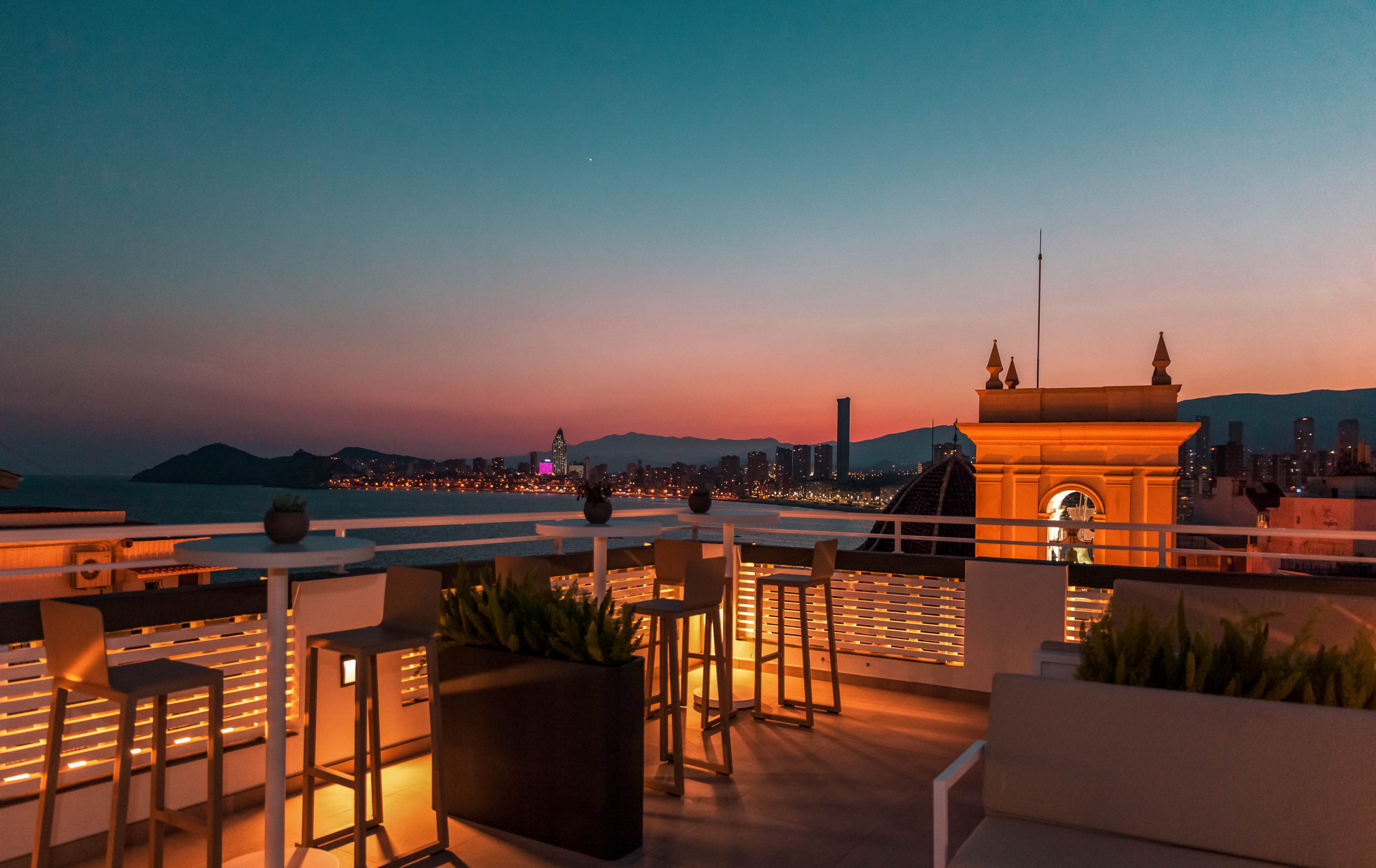 A modern rooftop terrace bar overlooks a coastal city at sunset. Tall bar stools and tables are arranged along the railing, with planters adding greenery to the warm-lit space. The sky transitions from deep blue to orange and pink, while distant buildings and mountains frame the horizon. Soft ambient lighting creates a relaxed, upscale atmosphere ideal for evening socializing.