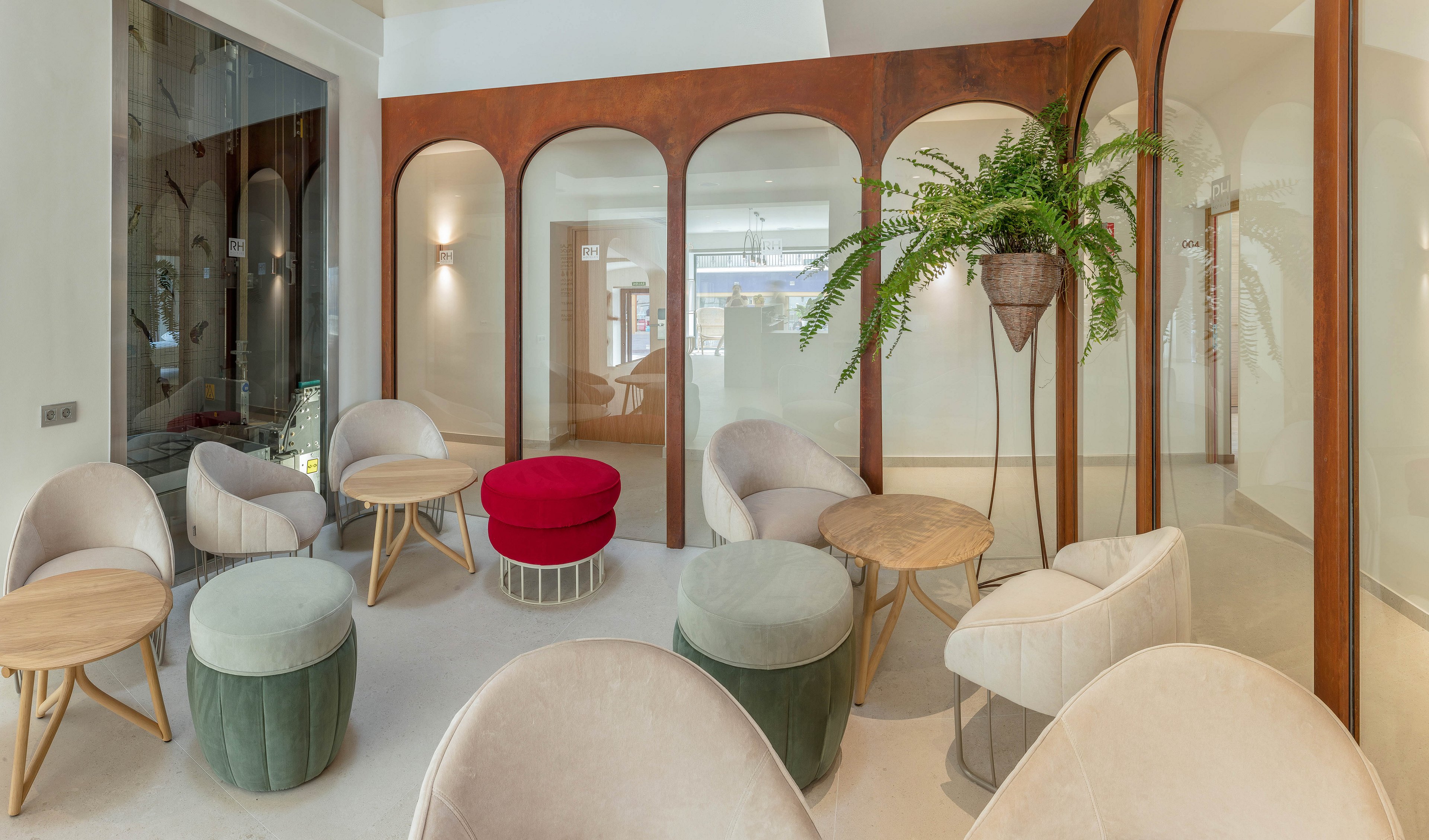 A contemporary lounge area features a mix of round chairs, stools, and small wooden tables arranged in a bright interior. Tall arched partitions with glass panels divide the space while maintaining an open, airy feel. Neutral beige tones are accented by a bold red ottoman and green upholstered stools. A hanging planter with lush greenery adds a natural touch to the modern design.