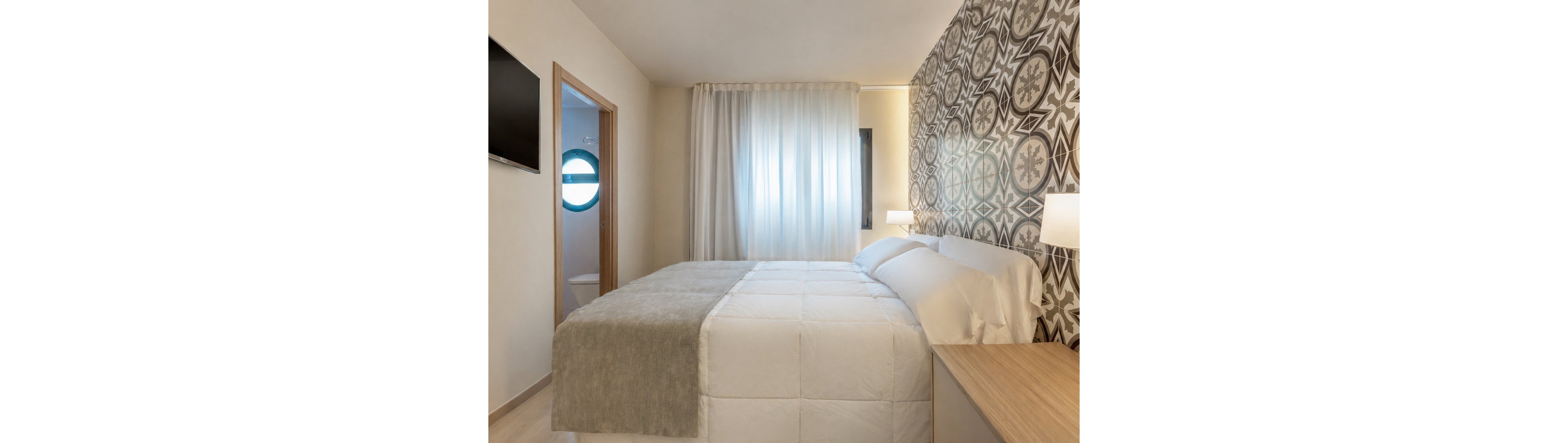A contemporary hotel bedroom features a neatly made double bed with white linens and a light gray throw blanket. One wall is decorated with bold geometric and floral patterned tiles, adding visual interest to the neutral space. Soft daylight filters through sheer curtains covering a large window, while a wooden nightstand and desk provide simple furnishings. A flat-screen television is mounted on the wall, and an open doorway reveals a glimpse of the adjoining bathroom.