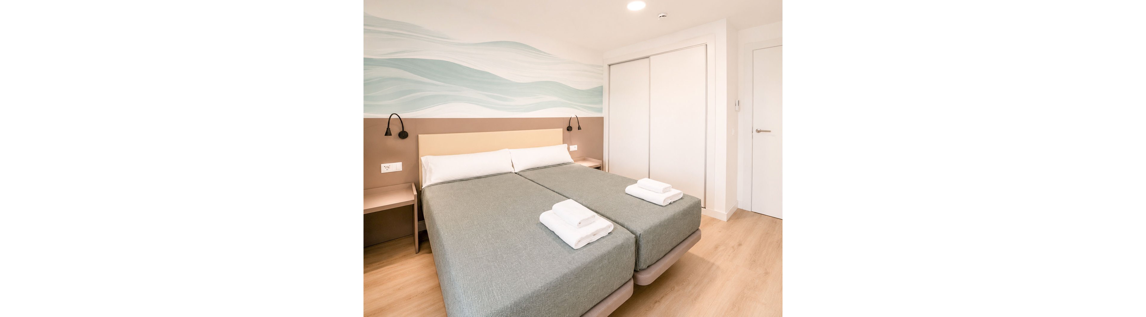 A bright, modern hotel room features two twin beds pushed together, each neatly topped with folded white towels. The space is decorated in soft neutral tones with a light wood floor and a minimalist wave mural on the wall behind the headboard. Built-in bedside tables with wall-mounted reading lights flank the bed, and a large white wardrobe with sliding doors stands opposite a plain white door. The overall atmosphere is clean, orderly, and inviting, suggesting a comfortable stay for guests.