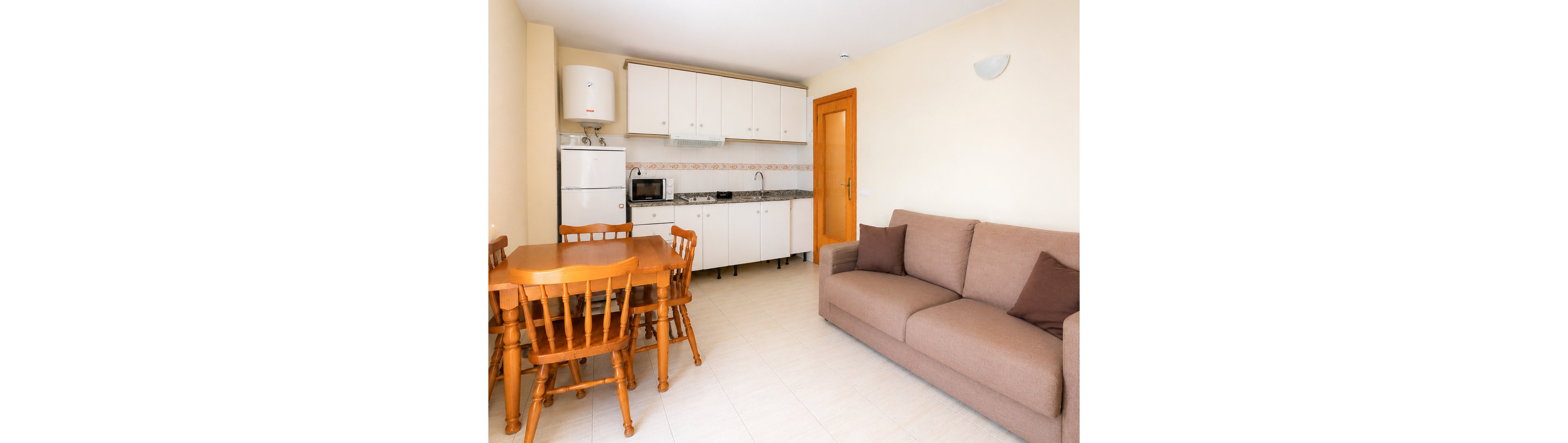 Bright studio apartment interior featuring a compact kitchenette and a comfortable sofa. The space includes white cabinets, a refrigerator, microwave, and sink along one wall. A wooden dining table with matching chairs sits near the kitchen area. Neutral tones and simple furnishings create a clean, functional living environment.