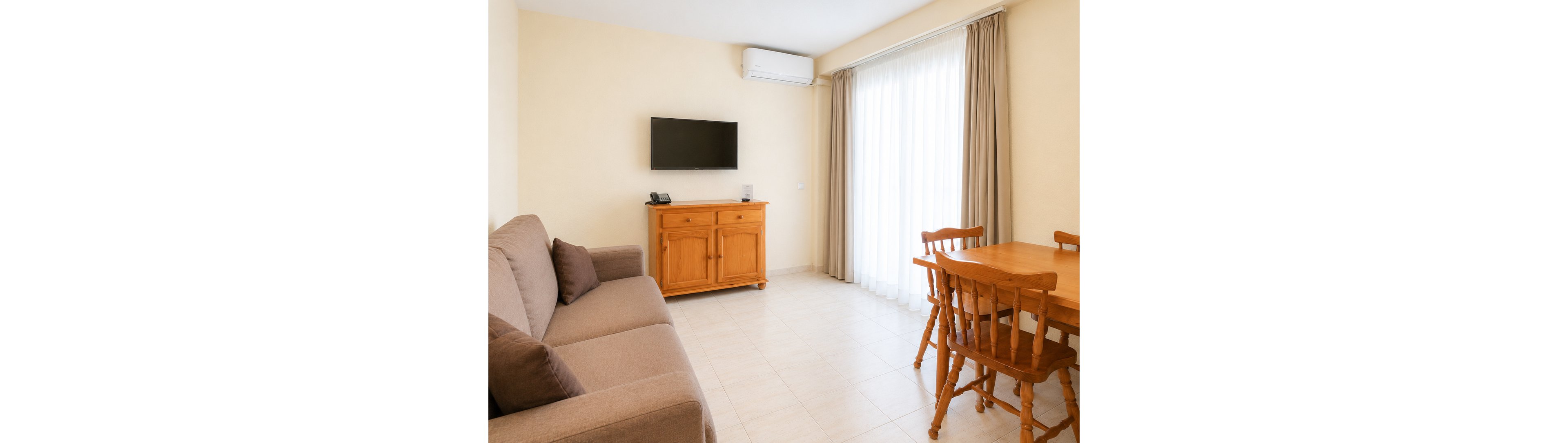 A cozy living room features a brown fabric sofa with cushions on the left side. A wooden cabinet stands against the wall, supporting a telephone and placed beneath a wall-mounted flat-screen TV. On the right, a wooden dining table with matching chairs sits near large floor-to-ceiling windows covered by sheer curtains and beige drapes. The space is lit by natural daylight, with light-colored tiled flooring and an air conditioning unit above the window creating a clean, comfortable atmosphere.